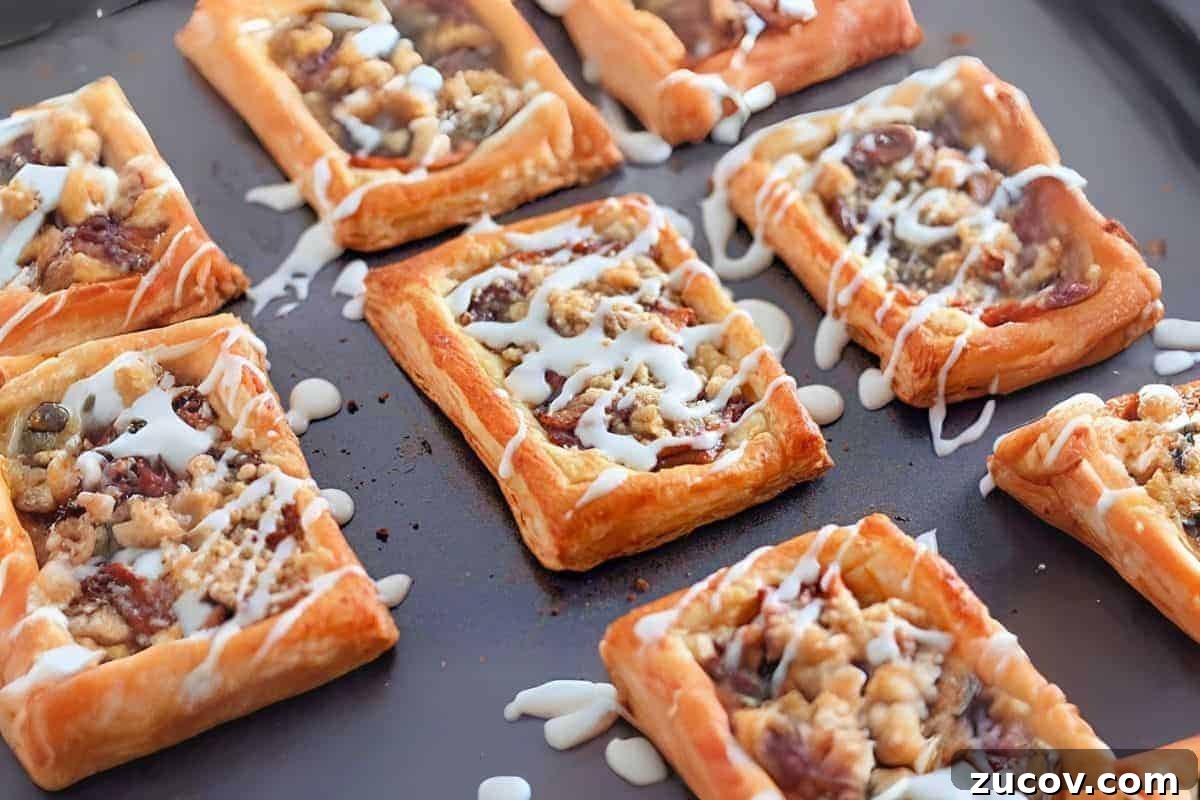 33 Irresistible Fall Desserts That Defy Portion Control 16 Golden brown Dutch Apple Danishes, topped with a fruit and nut filling, drizzled with white icing, displayed on a dark baking sheet.