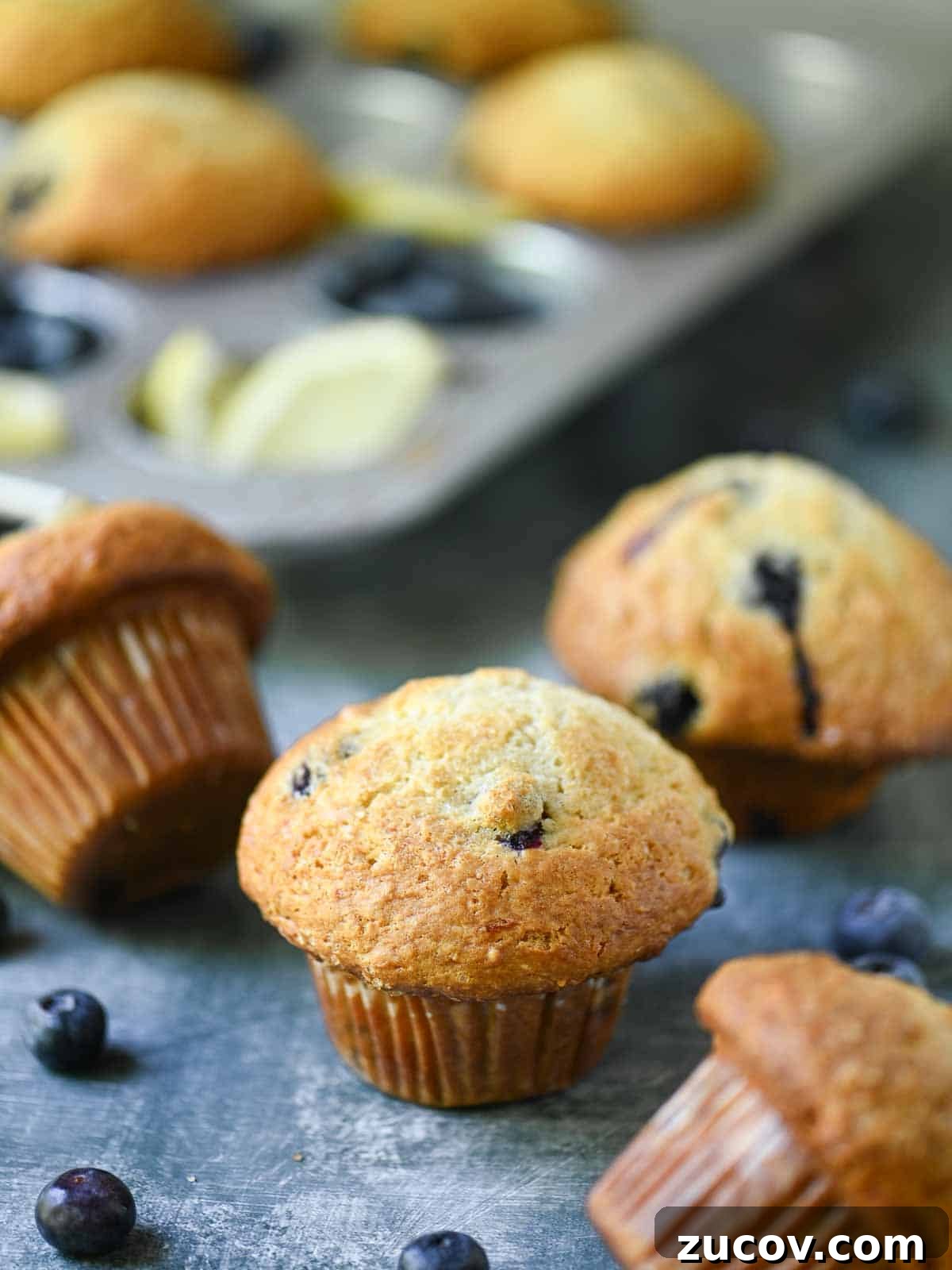 Bakery-Style Blueberry Lemon Muffins Recipe for Moist, Zesty Muffins 2 Scattered blueberry muffins in a muffin pan and on a blue surface.
