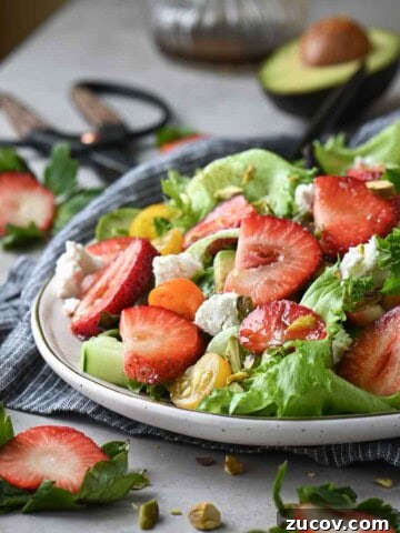 Close up of a strawberry salad with lettuce, tomatoes, goat cheese on a beige surface.