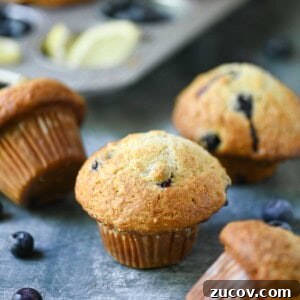 Bakery-Style Blueberry Lemon Muffins Recipe for Moist, Zesty Muffins 15 Scattered blueberry muffins in a muffin pan and on a blue surface.