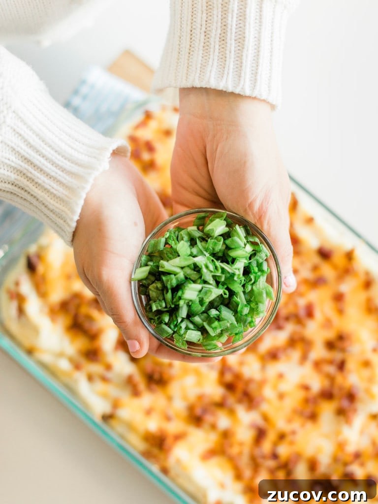 Creamy Loaded Mashed Potato Casserole with Bacon and Cheddar 6 Two hands holding bowl of chopped green onions above casserole in pan.