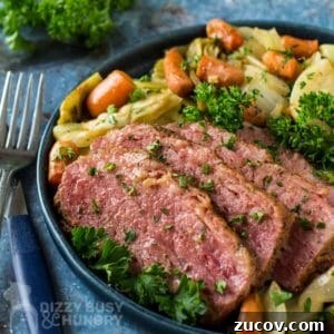 Irish Braised Corned Beef with Cabbage Recipe for St. Patrick’s Day 6 Side close up view of braised corned beef with vegetables garnished with herbs on a black plate with forks and herbs on the side on a blue cloth.