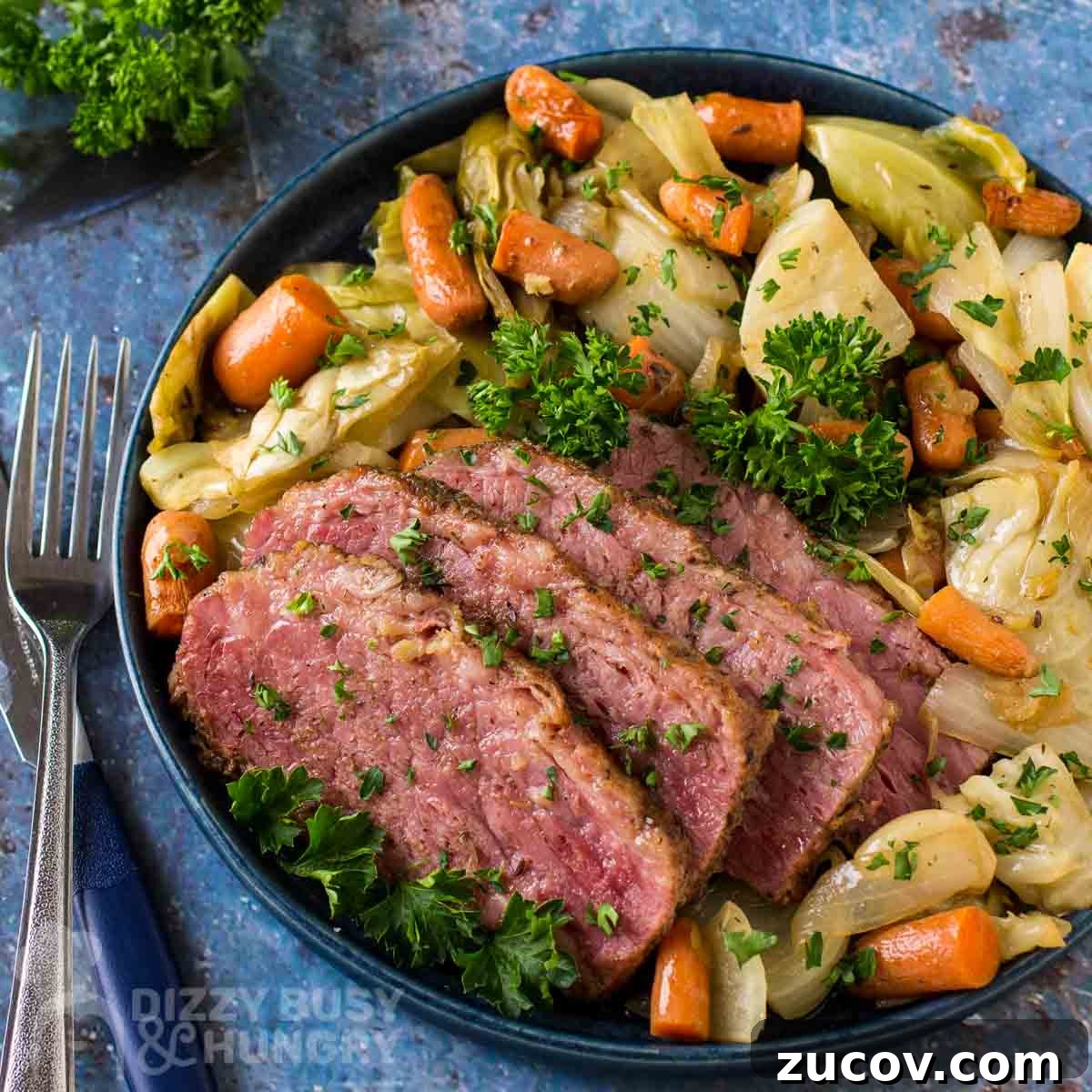 Irish Braised Corned Beef with Cabbage Recipe for St. Patrick’s Day 5 Overhead shot of braised corned beef with vegetables garnished with herbs on a black plate with forks and herbs on the side on a blue cloth.