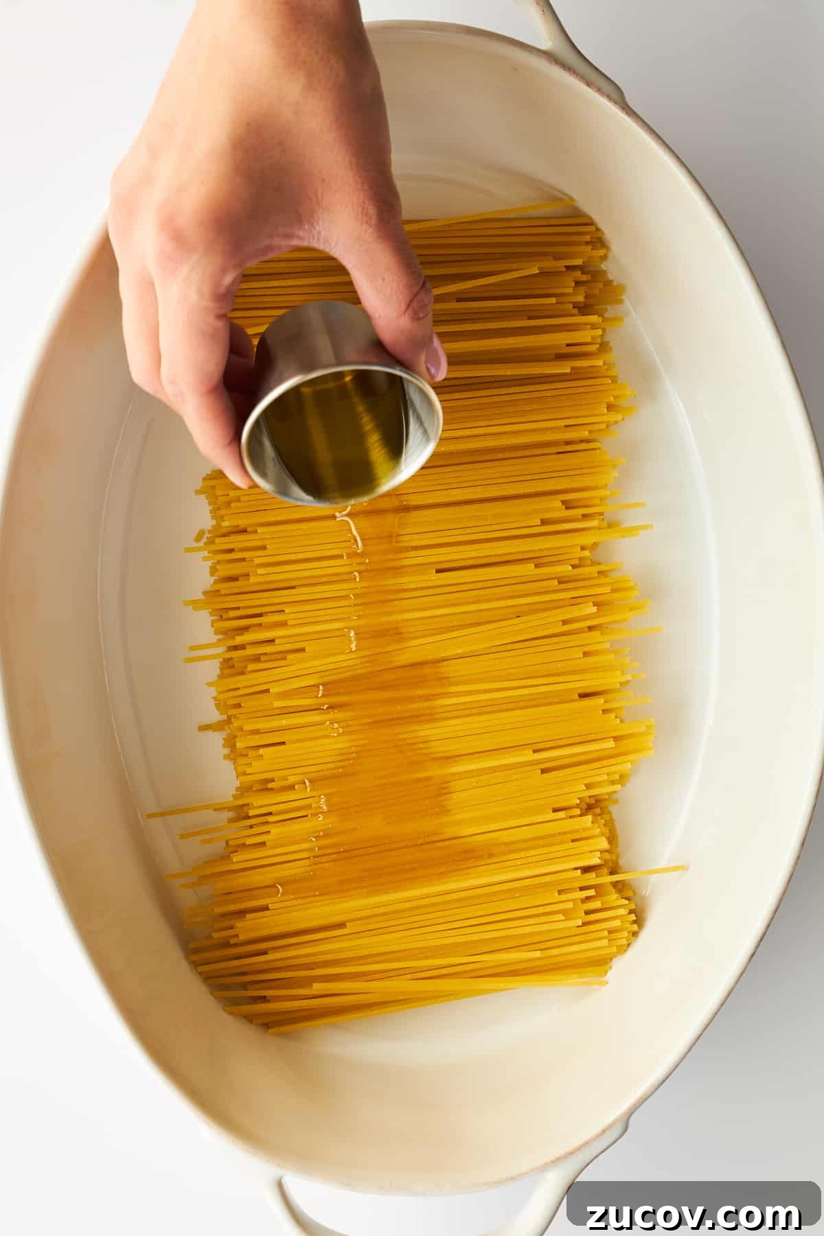 Cheesy Oven-Baked Spaghetti Casserole Recipe 4 Olive oil being poured over spaghetti noodles in an oval baking dish.