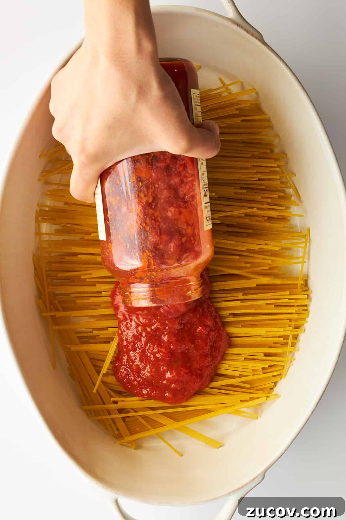 Cheesy Oven-Baked Spaghetti Casserole Recipe 5 Marinara sauce being poured over spaghetti noodles in an oval baking dish.