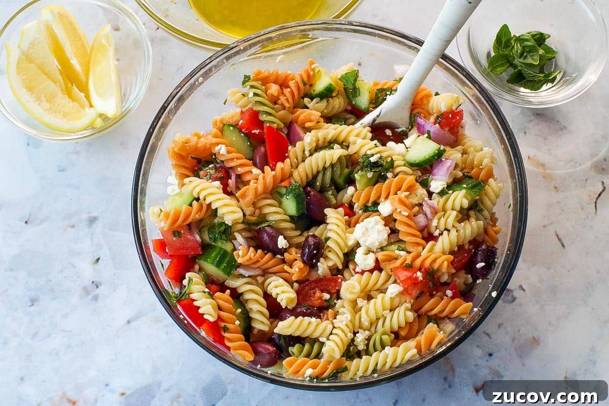 Greek Pasta Salad for Potlucks: Fresh Mediterranean Recipe 8 Remaining ingredients added to pasta in large glass bowl with spoon.