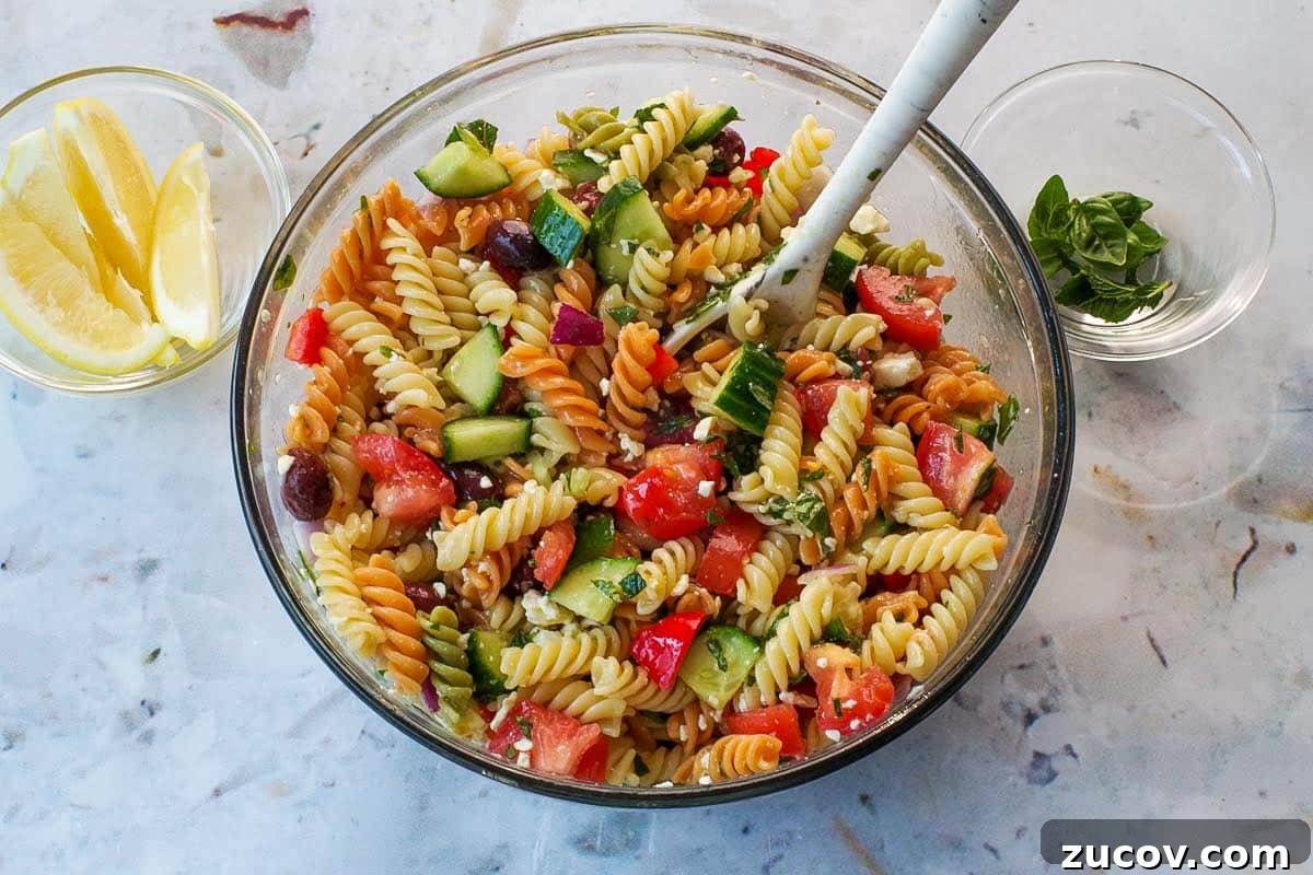 Greek Pasta Salad for Potlucks: Fresh Mediterranean Recipe 9 Salad dressing added to pasta and other ingredients in large glass bowl with spoon.
