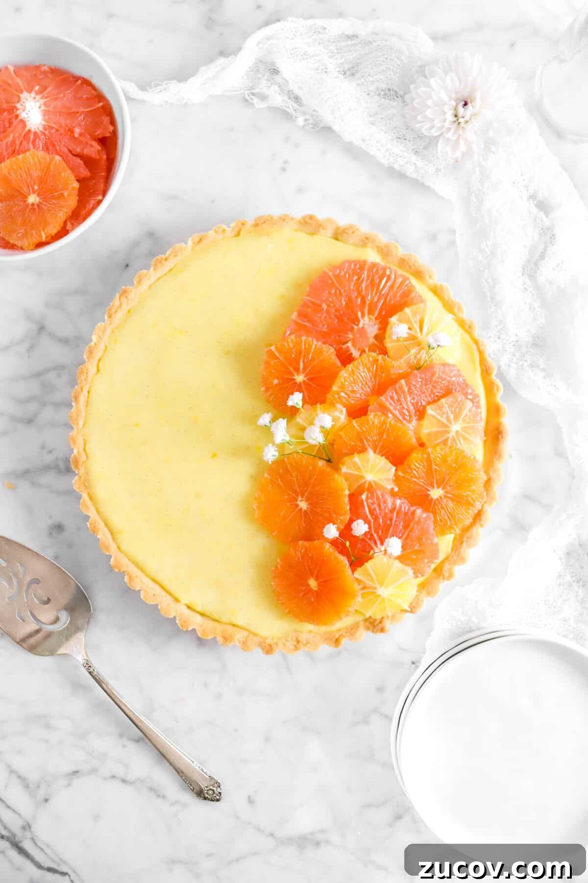 overhead shot of citrus tart with stacked plates, flowers, sliced citrus, a white napkin, and cake knife