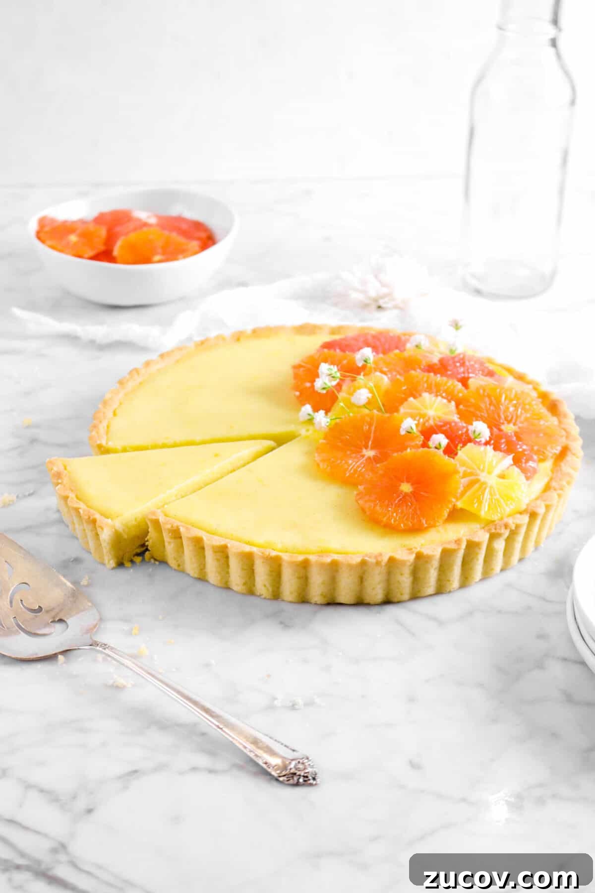 citrus tart with slice taken out of it on marble counter with sliced citrus, cake knife, and glass