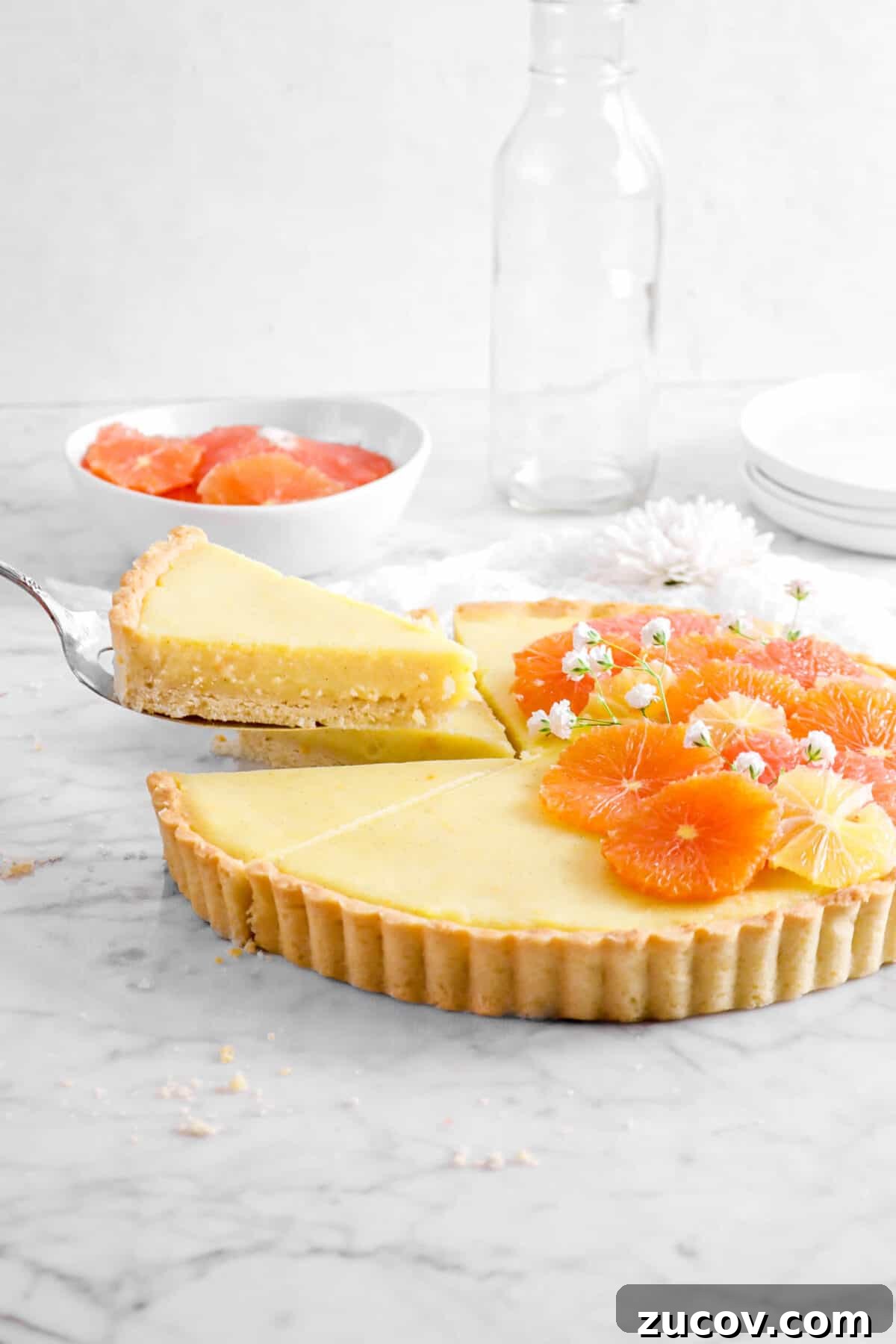 slice of citrus tart being held above the rest of the tart with bowl of citrus, glass, and stacked plates behind