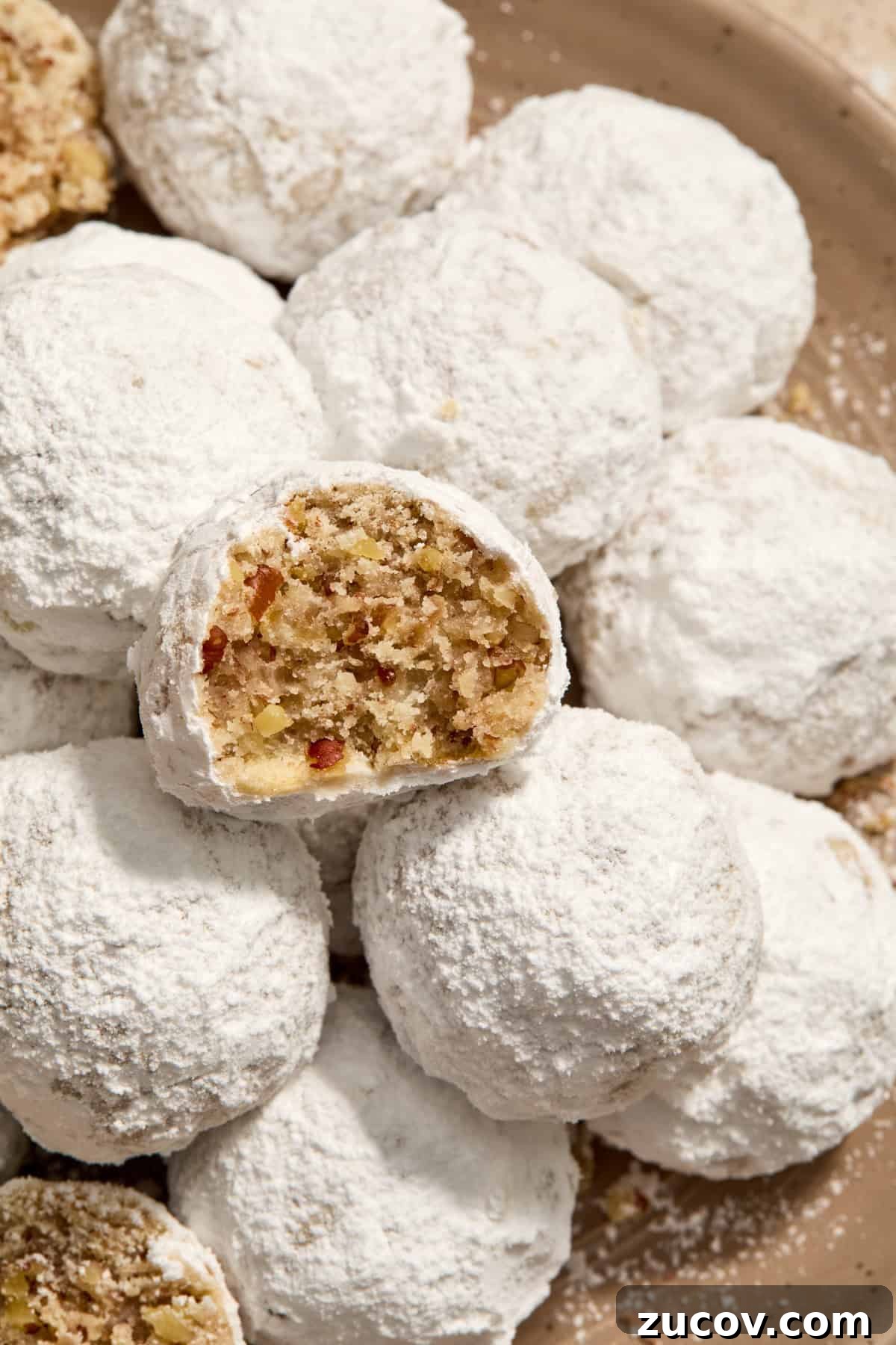 Cream Cheese Pecan Snowball Cookies for Christmas Baking 2 Cream cheese Christmas cookies with pecans and covered in powdered sugar on plate with bite taken out of one cookie.