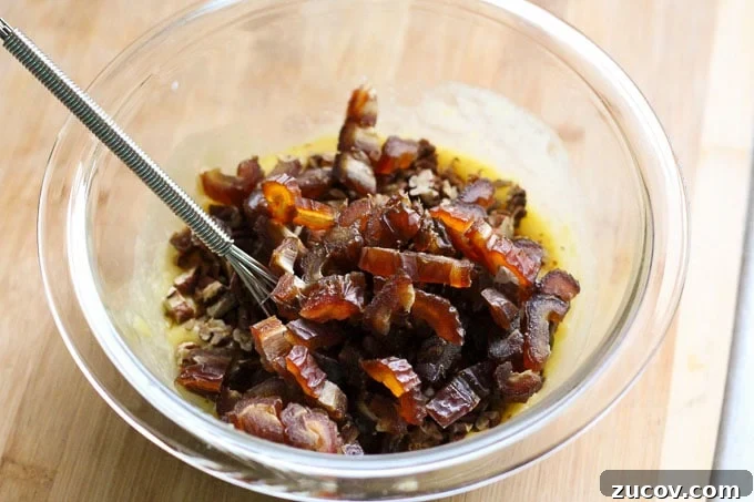 Paleo Baked Apples Recipe: Warm Spiced Fruit Dessert 3 A medium clear bowl with chopped up dates, nuts, lemon zest, and whisked egg in it. A small metal whisk is rested on the side of the bowl.