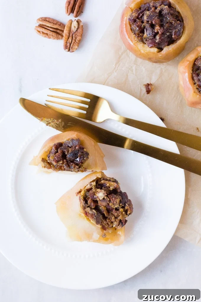 Paleo Baked Apples Recipe: Warm Spiced Fruit Dessert 4 a top down view of a white plate with a baked apple cut in half, gold knife and fork at the top, on top of parchment paper. In the upper left are three pecans and the upper right are two more baked apples