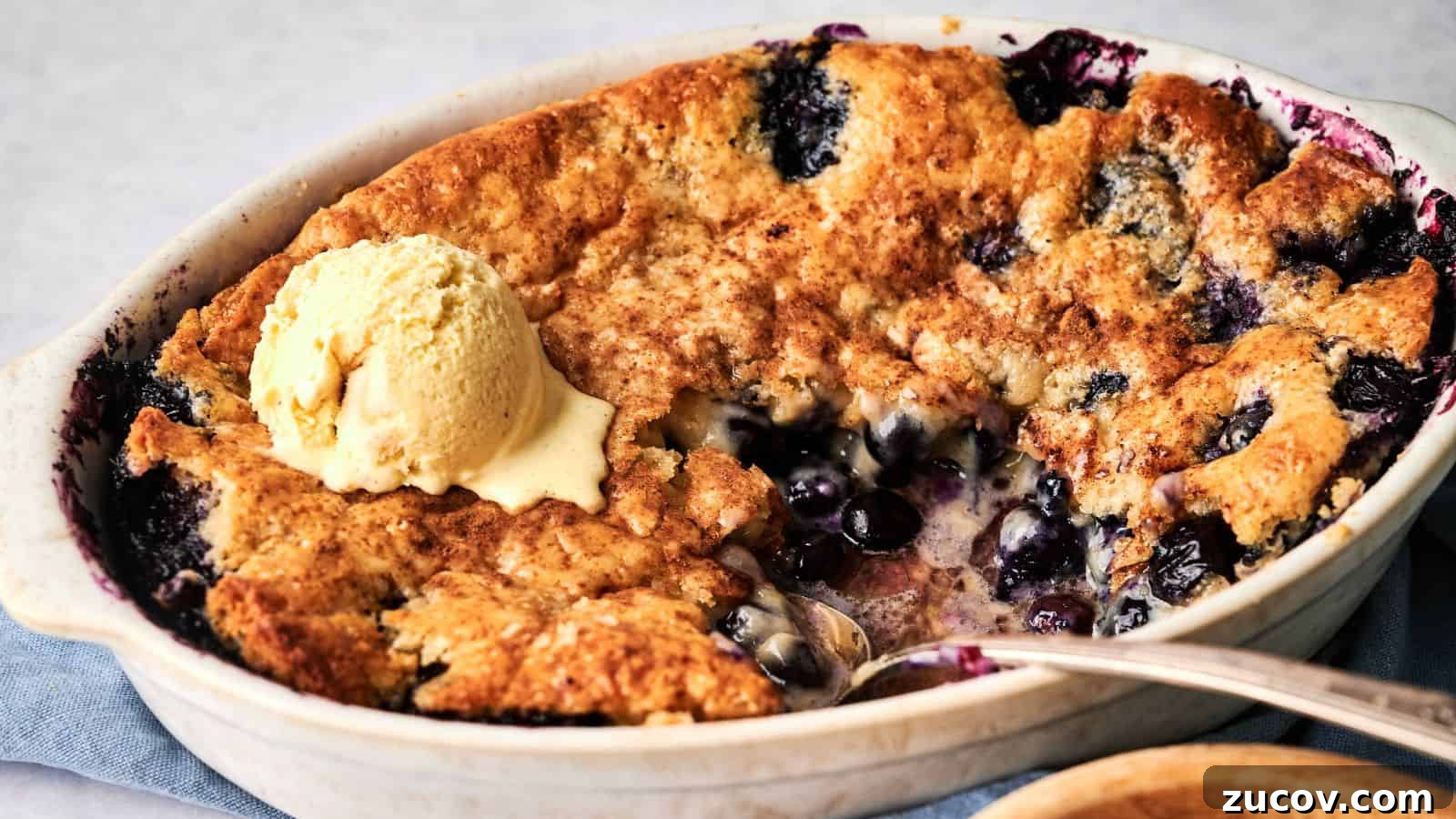 21 Summer Budget Meals That Make Takeout Obsolete 18 A baked blueberry cobbler with a golden crust is shown in a white dish, partially served, topped with a scoop of vanilla ice cream.