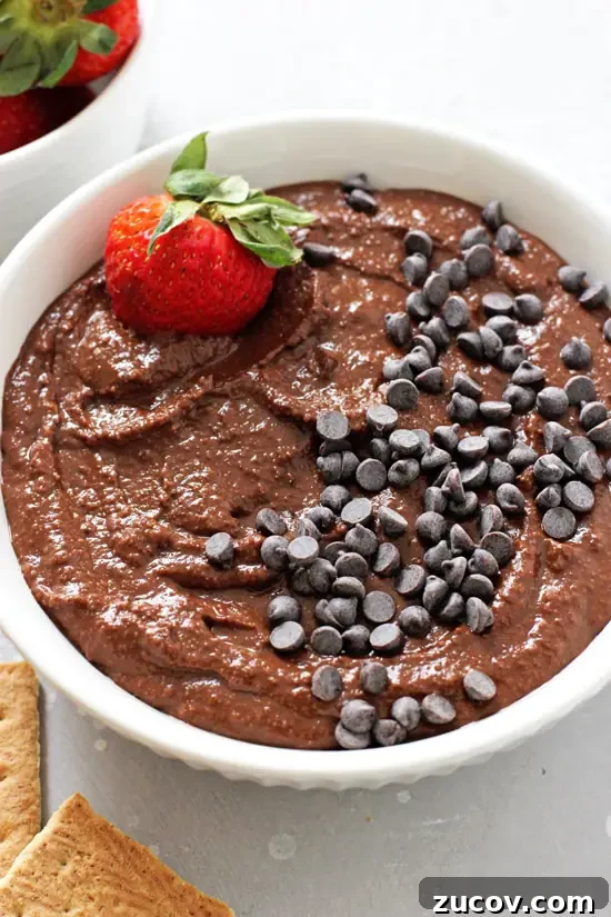 Easy and healthy, this chocolate hummus is addicting. Made with chickpeas, cocoa powder, almond butter and almond milk. A white bowl filled with Chocolate Hummus with a strawberry in the dish.
