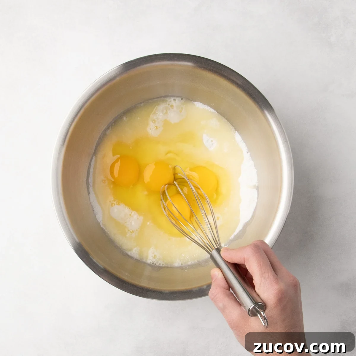 Gluten-Free Lemon Bar Recipe: Zesty, Tart Citrus Squares 13 Hand holding a metal whisk about to whisk together a mixture of white dry ingredients with eggs and lemon juice liquid mixture.