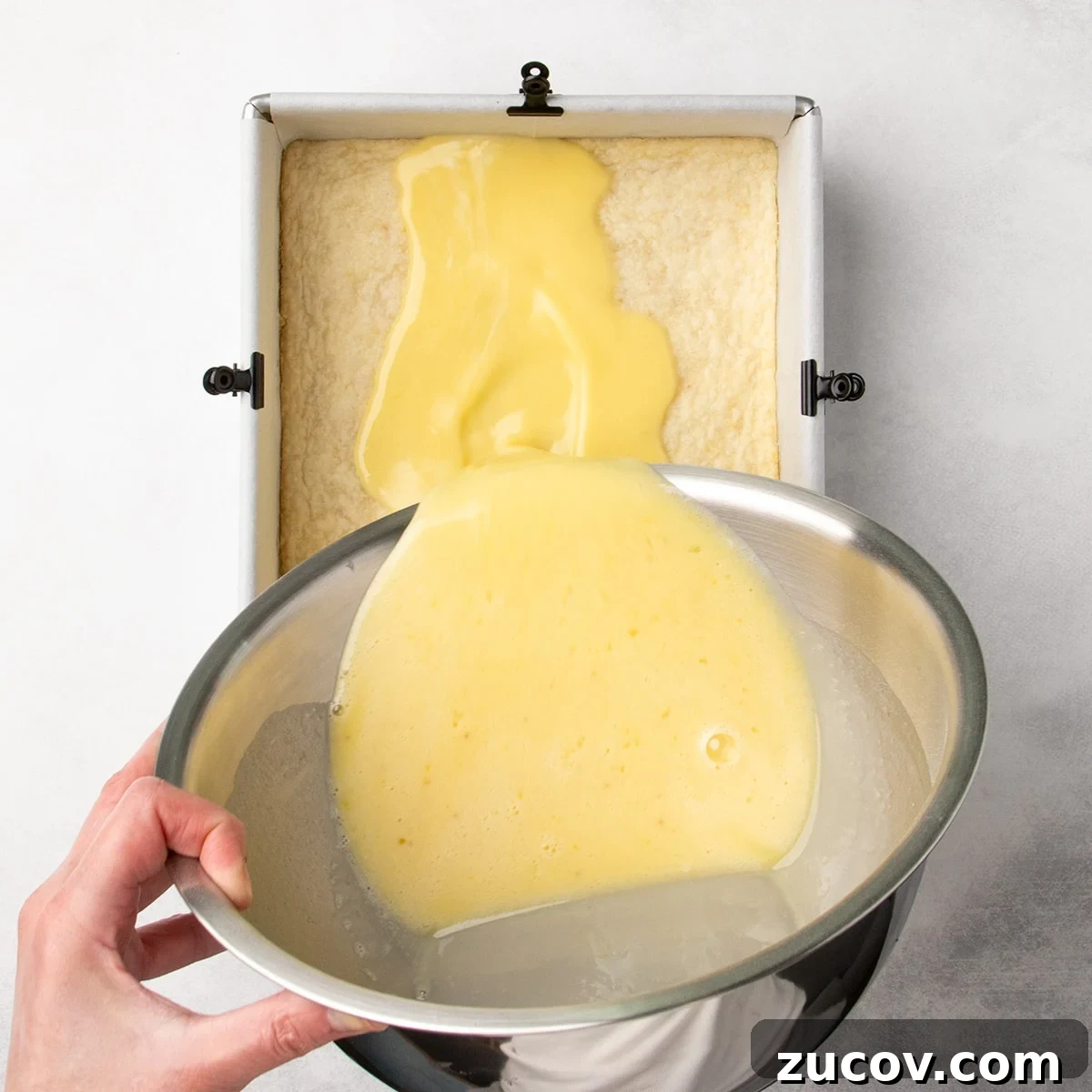 Gluten-Free Lemon Bar Recipe: Zesty, Tart Citrus Squares 14 Hand pouring a pale yellow liquid filling mixture from a metal bowl into a baked crust in a parchment-lined square pan.