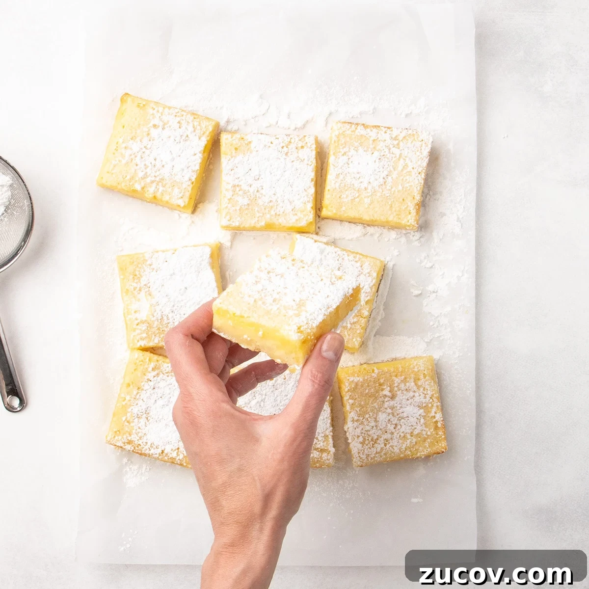 Gluten-Free Lemon Bar Recipe: Zesty, Tart Citrus Squares 17 Hand holding a square lemon bar dusted with powdered sugar over several cut bars on parchment paper, with a sieve partially visible to the side.