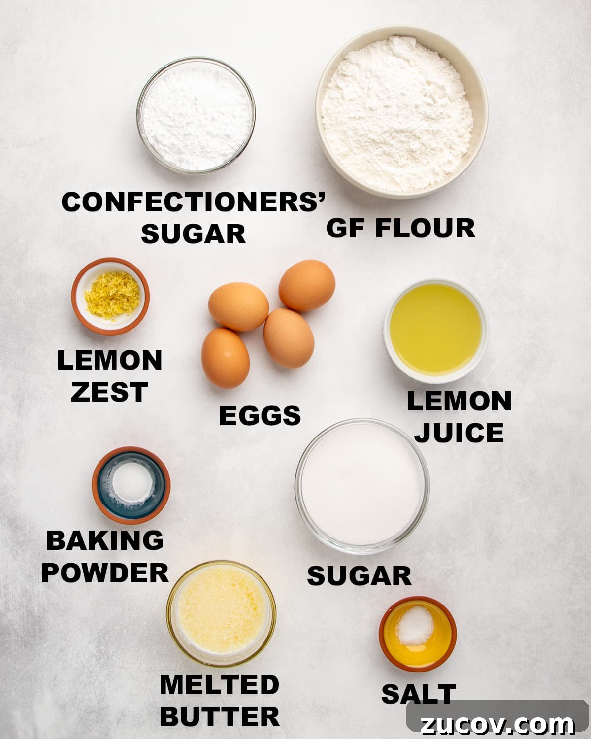 Gluten-Free Lemon Bar Recipe: Zesty, Tart Citrus Squares 3 Overhead view of measured ingredients in bowls with labels including confectioners’ sugar, gluten free flour, lemon zest, eggs, lemon juice, baking powder, sugar, melted butter, and salt.