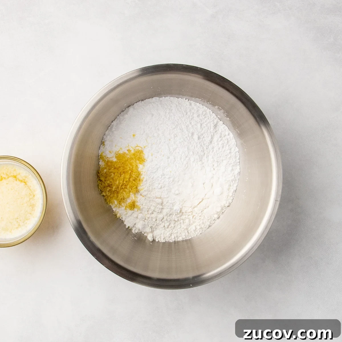 Gluten-Free Lemon Bar Recipe: Zesty, Tart Citrus Squares 4 Metal mixing bowl filled with white dry ingredients and a small pile of yellow lemon zest, with a glass of melted butter mixture partially visible to the side.