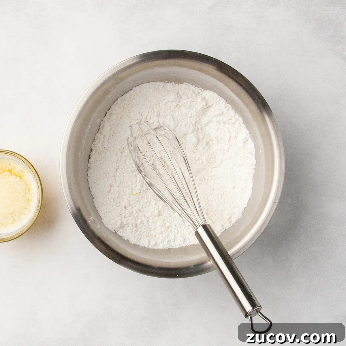 Gluten-Free Lemon Bar Recipe: Zesty, Tart Citrus Squares 5 Metal mixing bowl with white dry ingredients being whisked with a metal whisk, with some visible yellow flecks of lemon zest