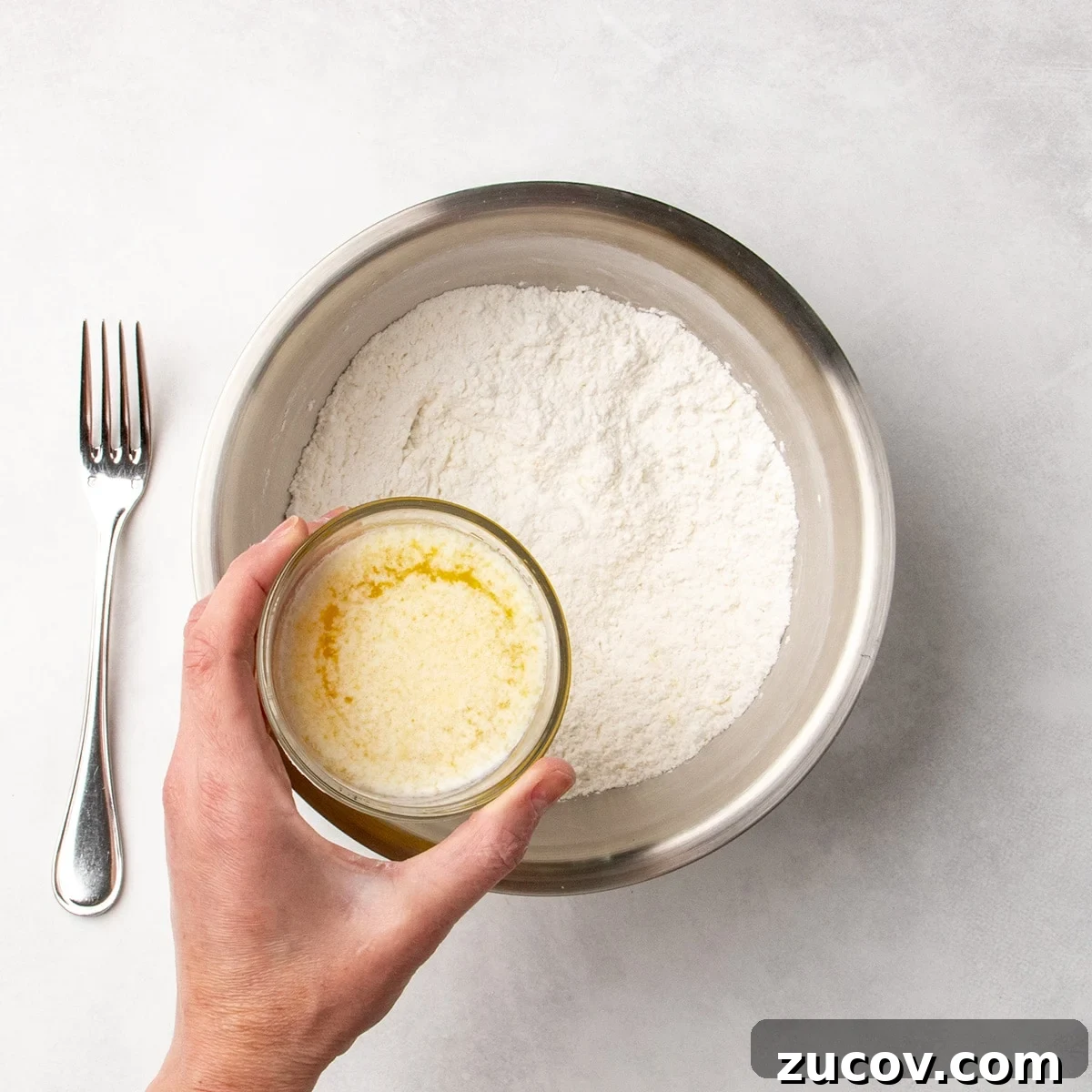 Gluten-Free Lemon Bar Recipe: Zesty, Tart Citrus Squares 6 Hand holding a small glass of melted butter mixture about to pour it into a bowl of white dry ingredients, with a fork resting nearby.
