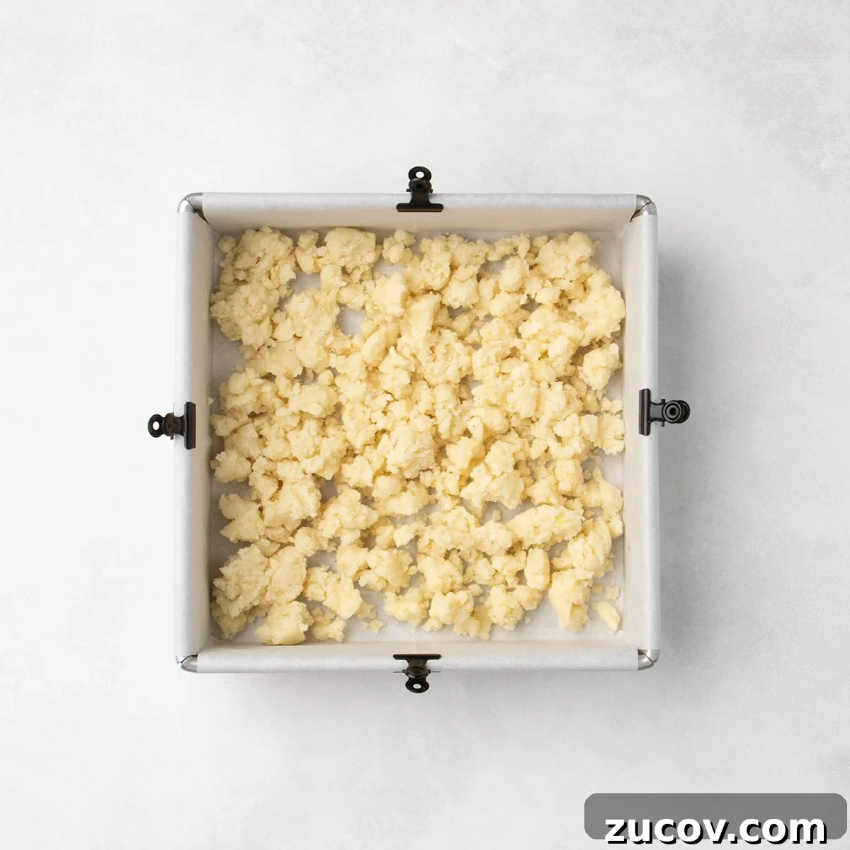 Gluten-Free Lemon Bar Recipe: Zesty, Tart Citrus Squares 9 Pale dough clumps scattered in an even layer in a square baking pan lined with parchment paper, clipped to each of the four sides with a small black binder clip.