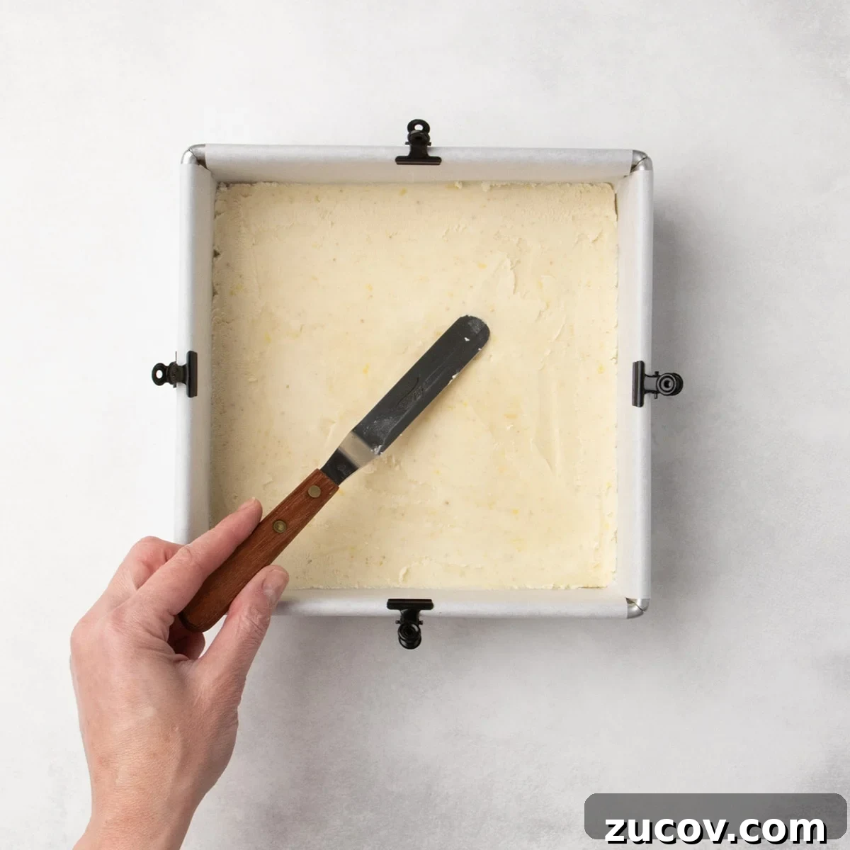 Gluten-Free Lemon Bar Recipe: Zesty, Tart Citrus Squares 10 Hand holding a small offset spatula to press pale dough into a smooth, even layer in a parchment-lined square pan.