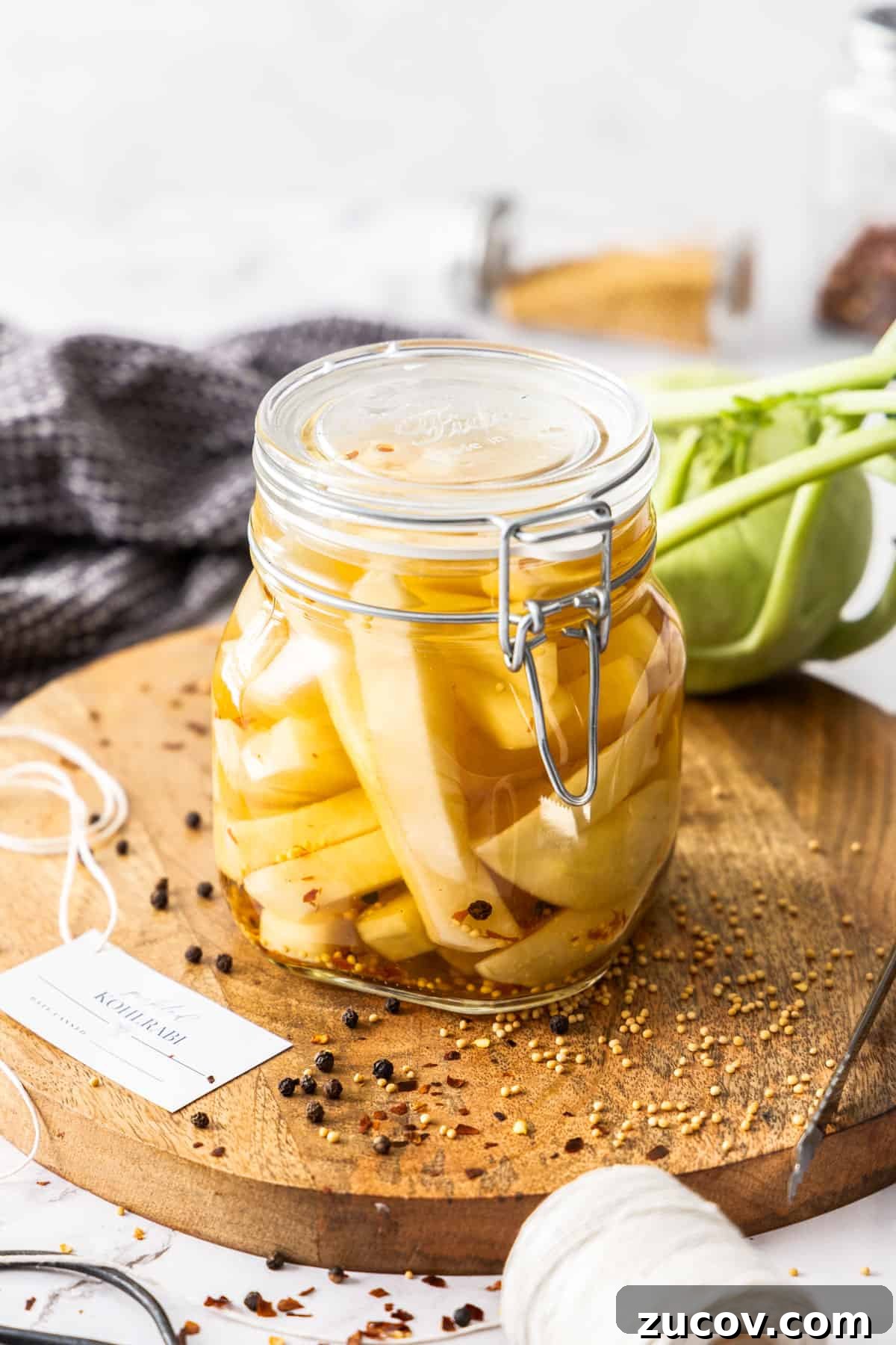 Crispy Pickled Kohlrabi Recipe for Tangy, Quick Ferments 7 Jar of Pickled Kohlrabi, sitting on a round wooden board, with some herbs and spices around the edge.