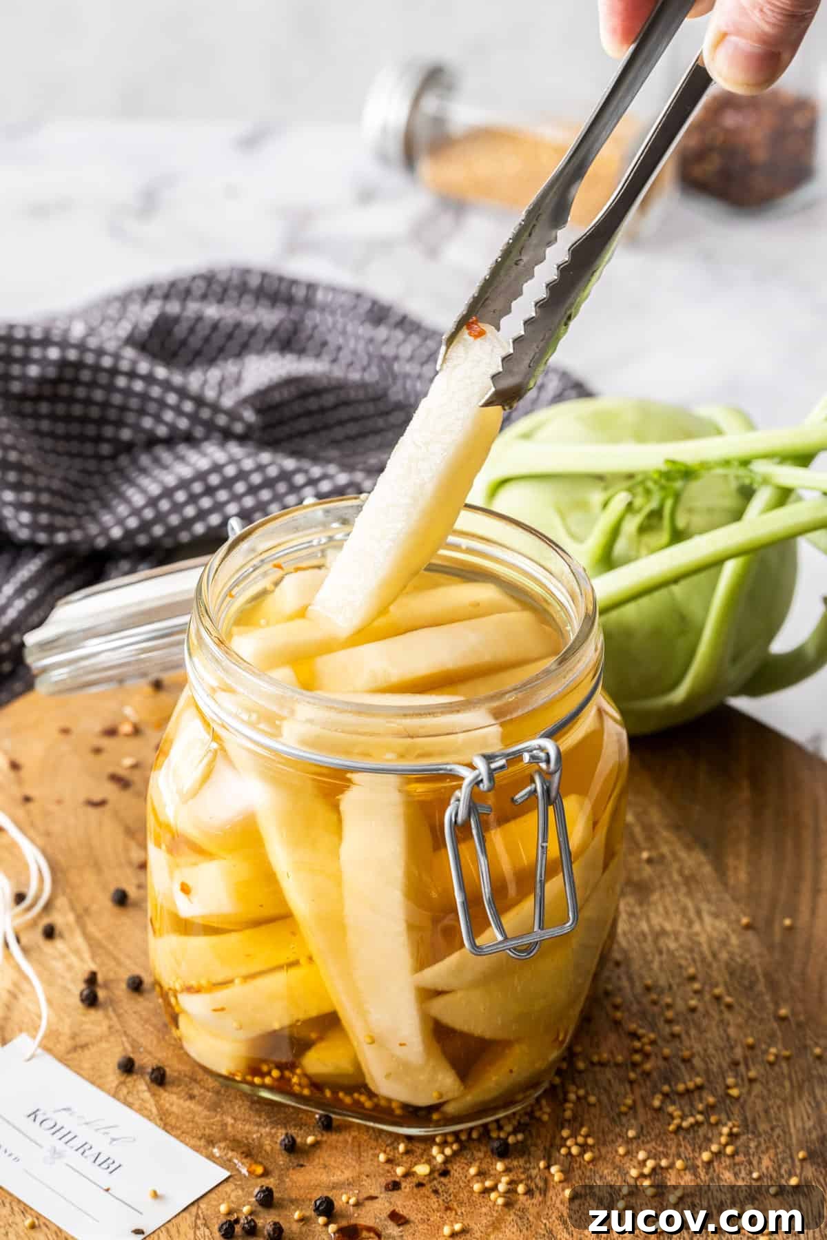 Crispy Pickled Kohlrabi Recipe for Tangy, Quick Ferments 8 Jar of Pickled Kohlrabi, sitting on a round wooden board, with some tongs reaching in to pick up a piece of kohlrabi.