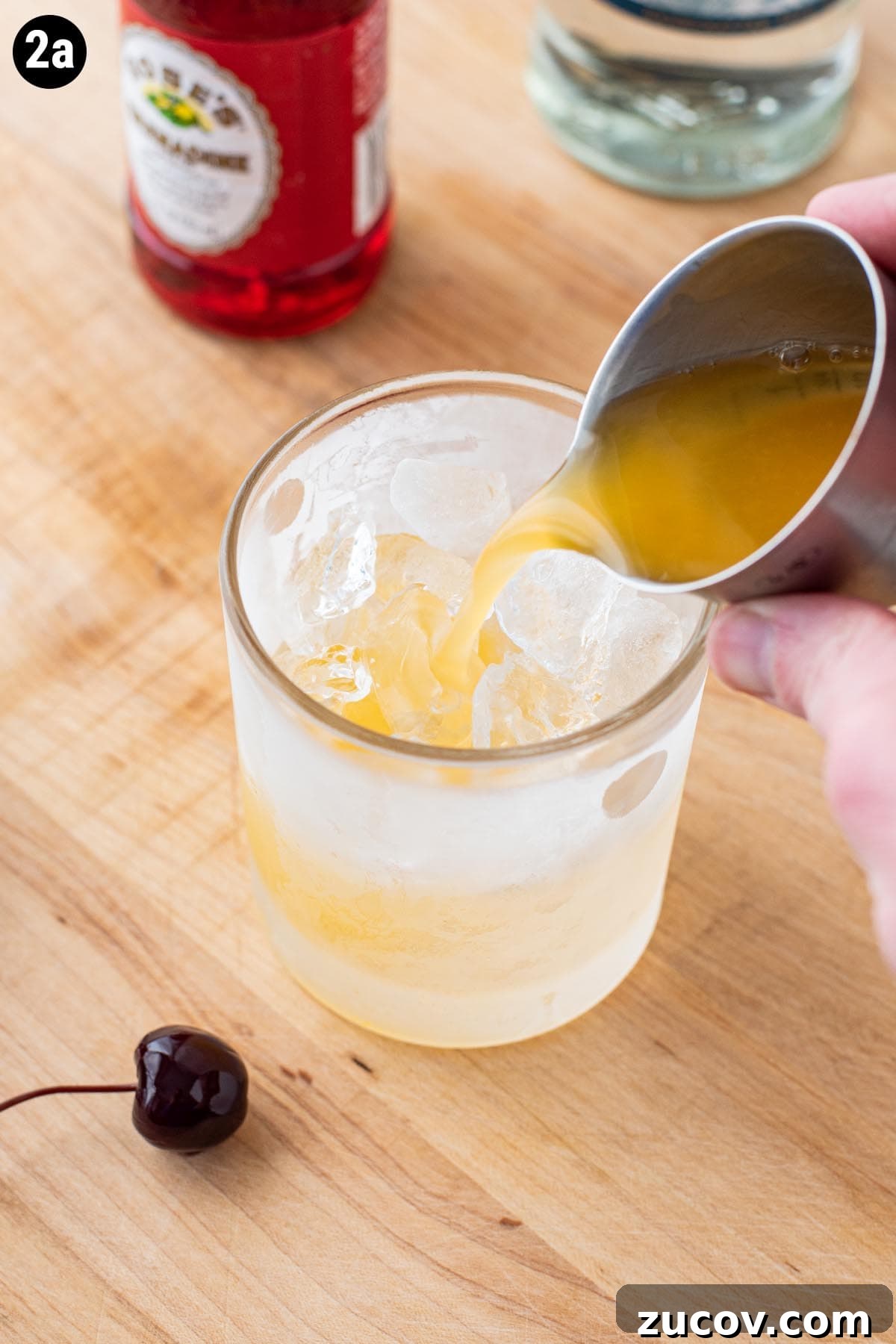 Tequila Sunrise Cocktail Recipe: Ingredients, Tips & Variations 6 A hand holding a jigger pouring fresh orange juice into an ice filled double old fashioned glass.