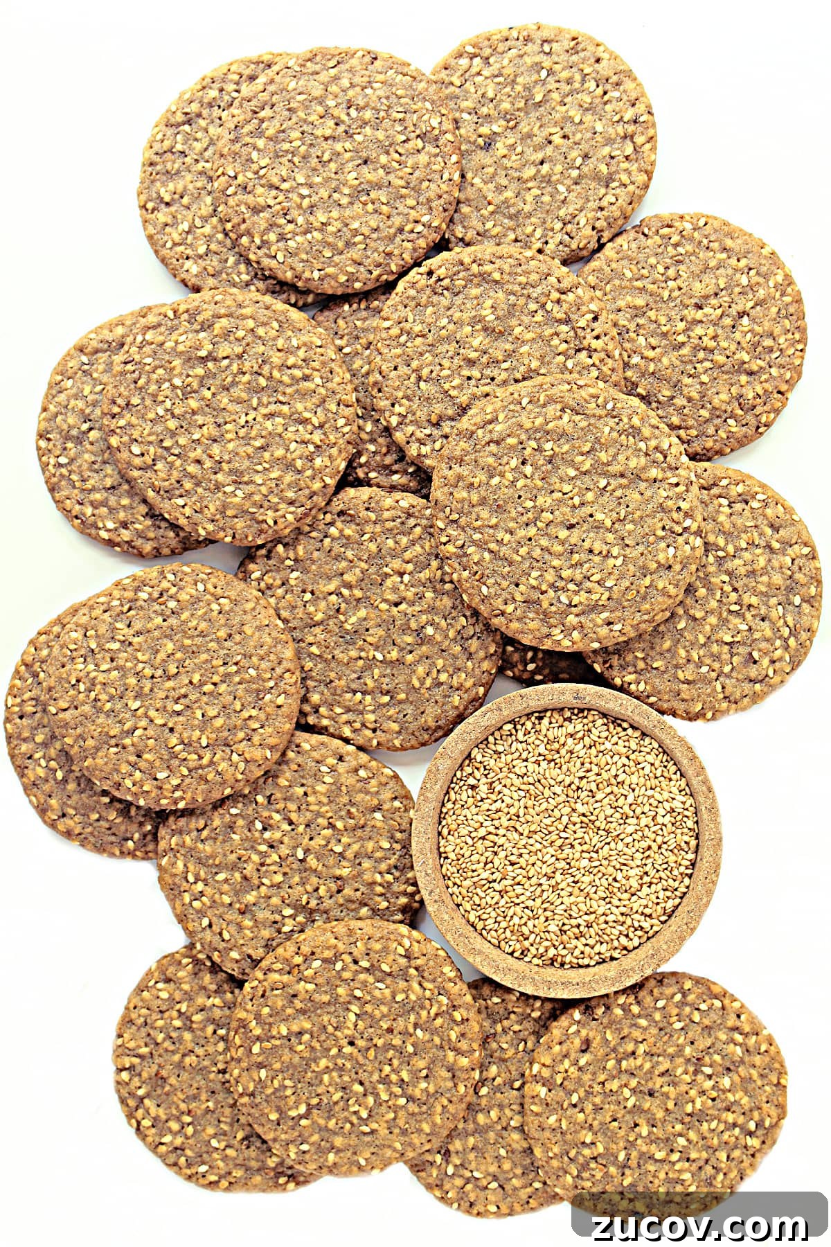 Sesame Benne Wafers: Crispy South Carolina Cookie Recipe 2 Brown sesame wafer cookies with a bowl of toasted sesame seeds on a white background.