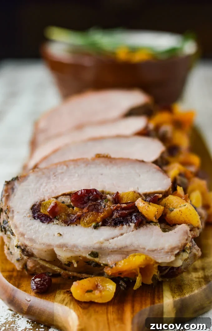 Fruit-Stuffed Pork Loin Roast with Apple and Cranberry Filling 9 Slices of cooked fruit stuffed pork loin roast sitting on a wooden cutting board with a bowl of fruit sauce sitting in the background.