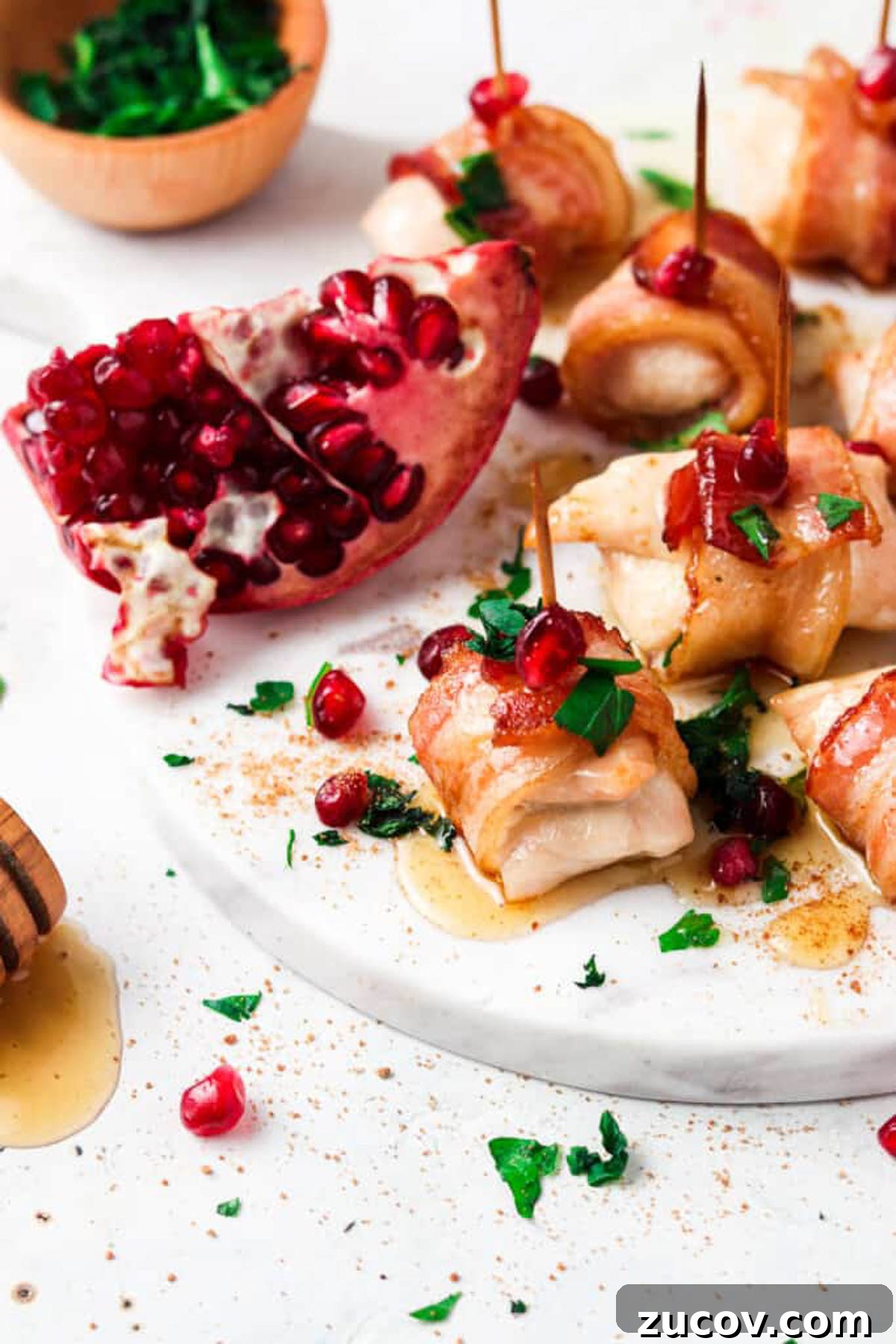 Crispy Bacon-Wrapped Chicken Bites with Maple Glaze 5 Bacon wrapped chicken bites drizzled with honey and garnished with fresh green herbs and pomegranate seeds on a marble platter.