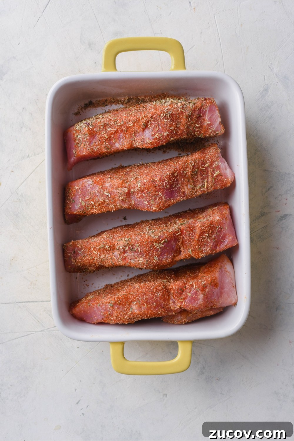 Oven-Baked Boneless Country-Style Pork Ribs — Juicy and Tender 6 Overhead view of four seasoned and boneless ribs placed evenly in a white baking dish with yellow handles.