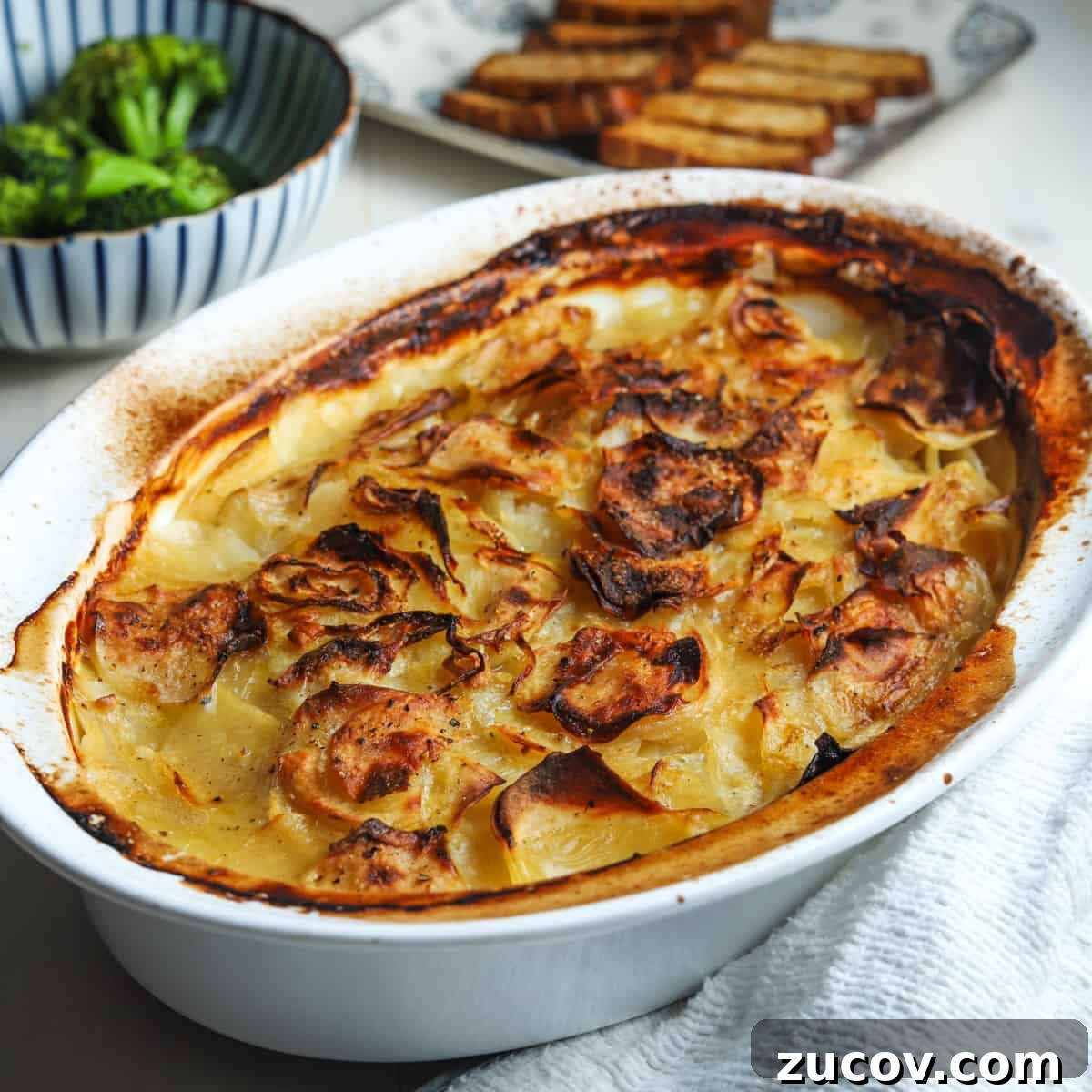 Photo of vegan boulangere potatoes in a dish with dishes of broccoli and smoked tofu slices int eh background.