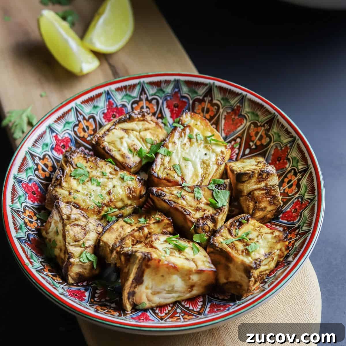 Photo of roast celeriac in a colourful bowl on a wooden board with lemon wedges, fresh herbs and a black background.
