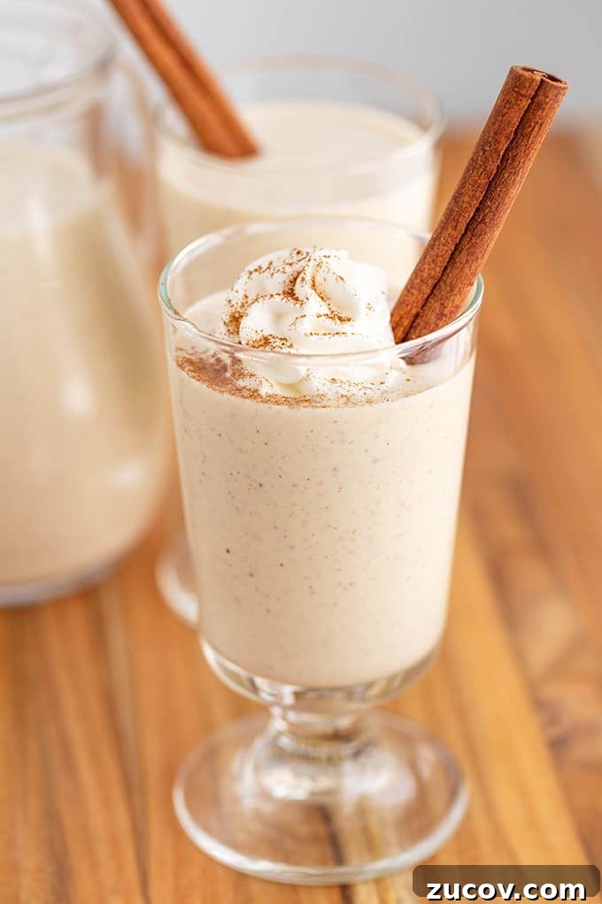 Classic Eggnog Recipe: Spiked and Alcohol-Free Versions 4 Eggnog in a glass with whipped cream, cinnamon stick, and a sprinkle of cinnamon on a wood surface