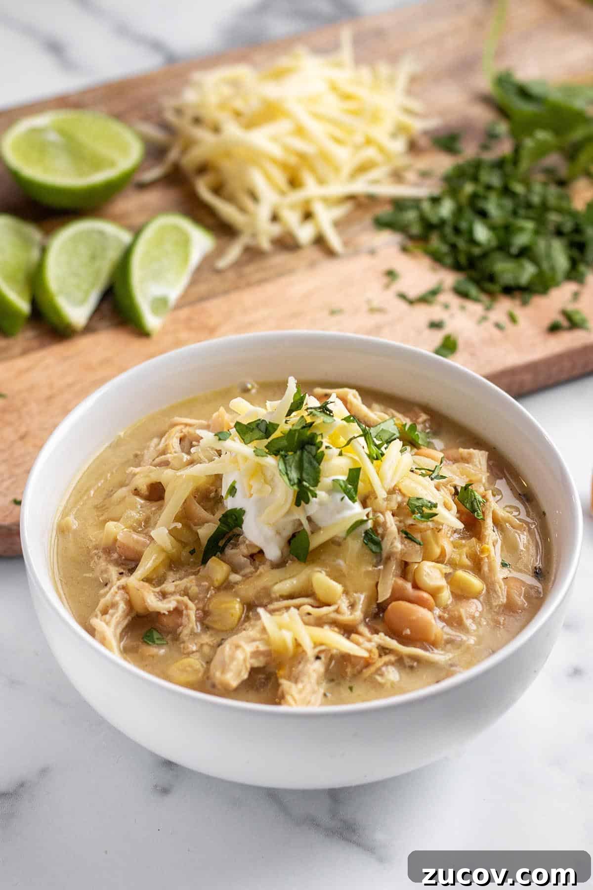Slow-Cooker Creamy White Chicken Chili Recipe 2 Bowl with white chicken chili soup topped with sour cream, cilantro and cheese. In the back is limes, shredded cheese and cilantro on a board.