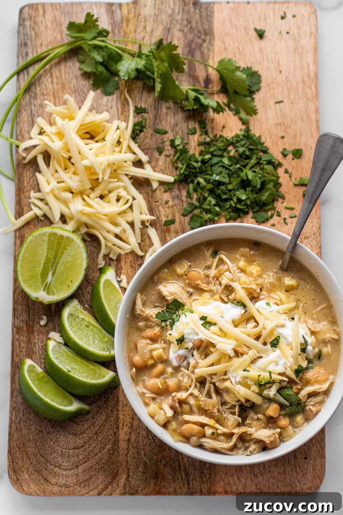 Slow-Cooker Creamy White Chicken Chili Recipe 13 Serving board with a bowl of white chicken chili, limes, shredded cheese and cilantro.