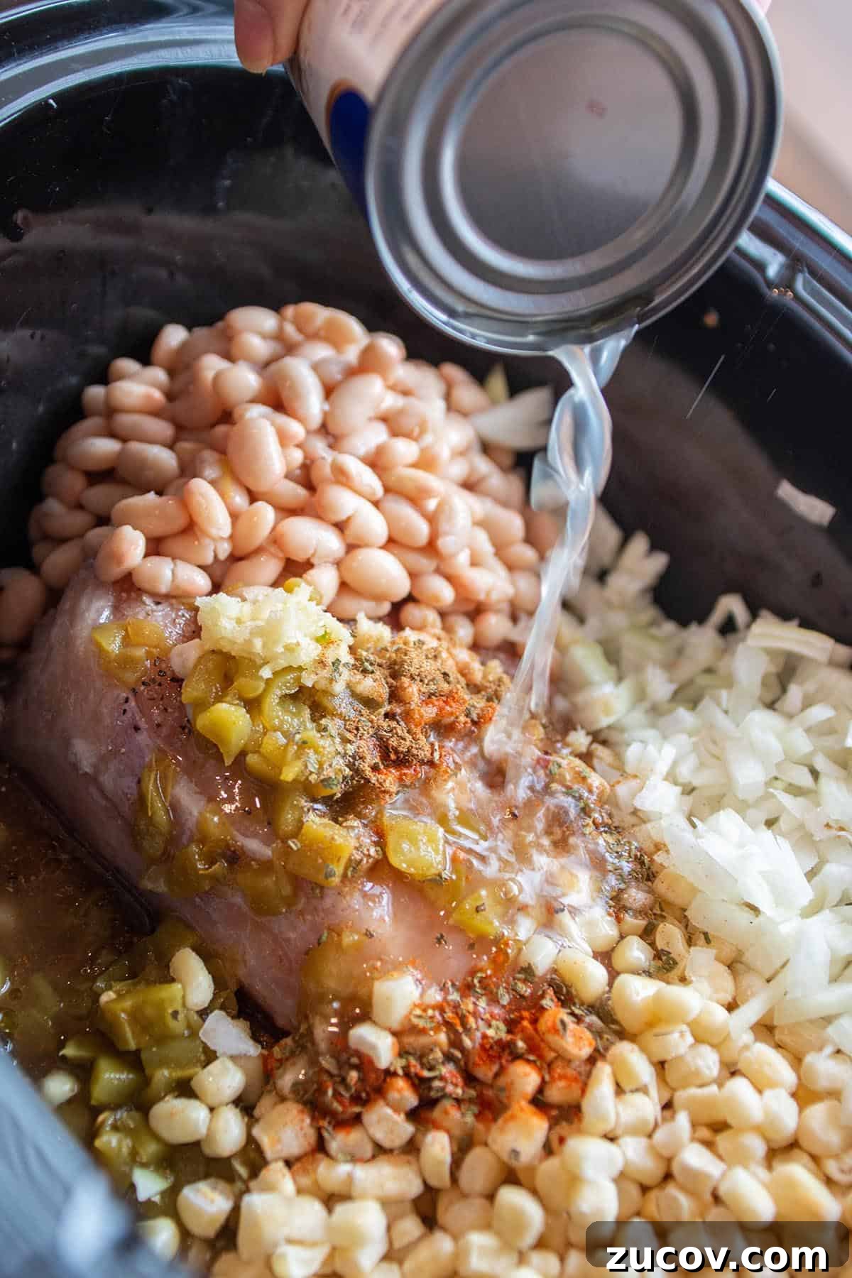 Slow-Cooker Creamy White Chicken Chili Recipe 5 Crock-pot full of fresh onions, white beans, corn, frozen chicken and spices with chicken broth being poured in.