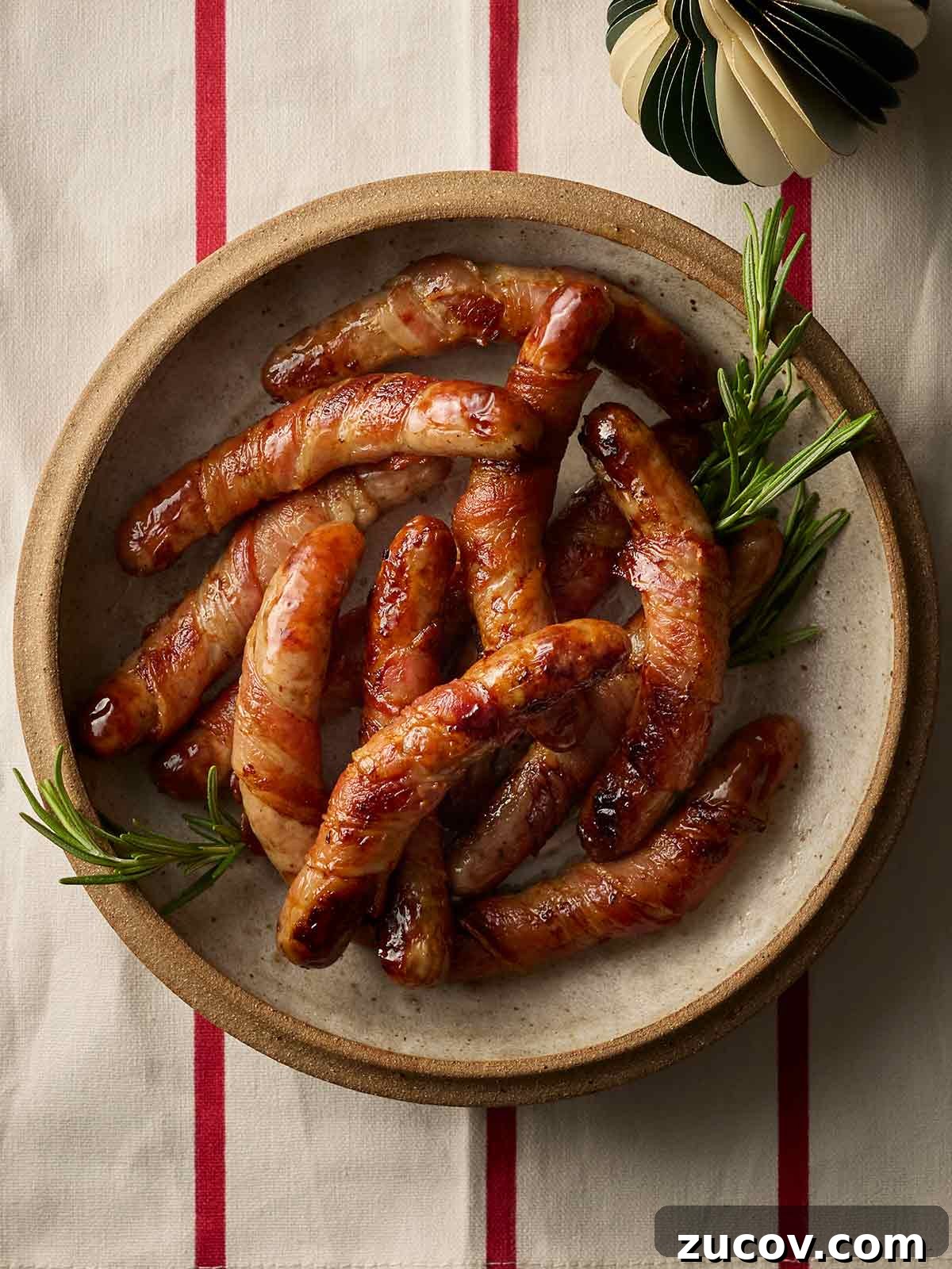 Savory Sausage-Wrapped Puff Pastry Bites for Parties 2 A plate of cooked Pigs in Blankets with sprigs of rosemary, on a red and white stripy tablecloth.