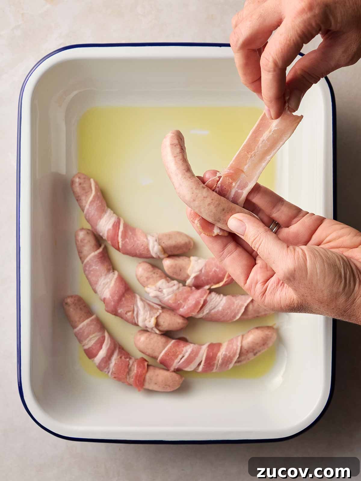 Savory Sausage-Wrapped Puff Pastry Bites for Parties 6 A white oven dish with oil in and sausages wrapped with bacon around, and a hand wrapping up a raw Pig in Blanket.