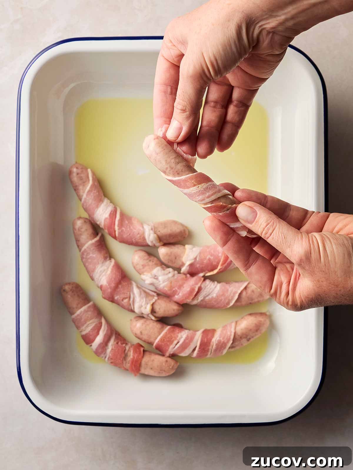 Savory Sausage-Wrapped Puff Pastry Bites for Parties 7 Two hands wrapping bacon around sausages with an oven dish ready to cook Pigs in Blankets.