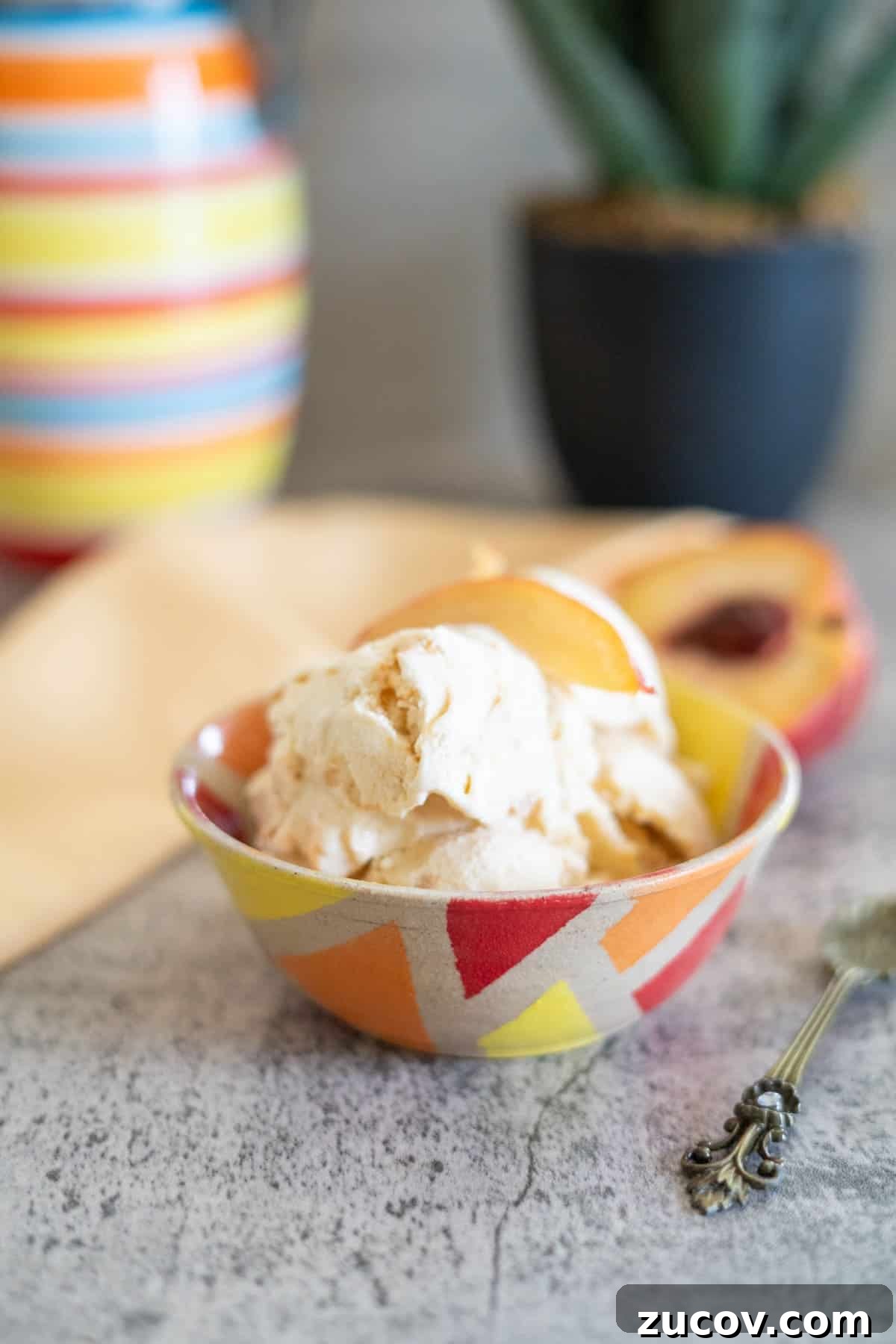 Keto Peach Ice Cream Recipe: Creamy Low-Carb Frozen Dessert 2 A bowl of ice cream with peach slices, next to a decorative spoon and a striped vase, on a gray countertop.