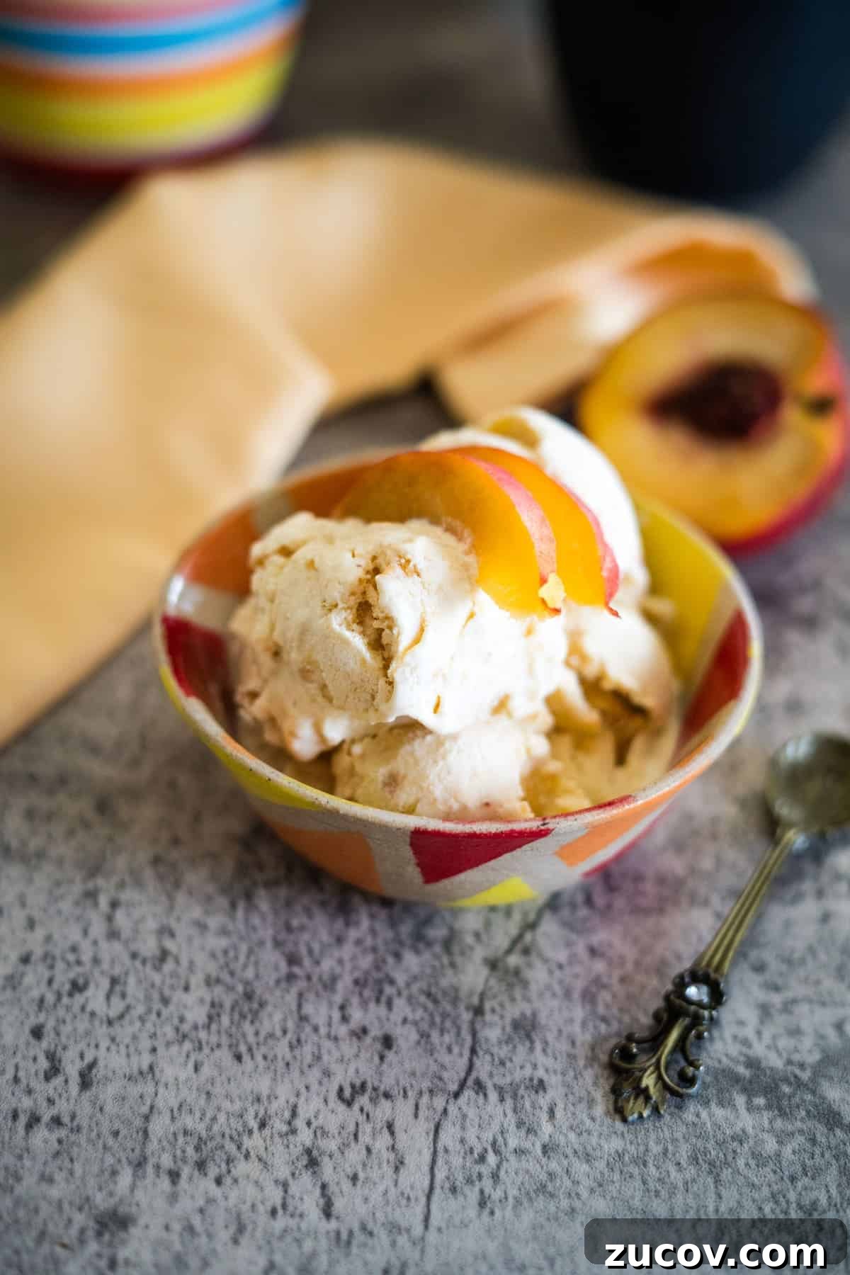 Keto Peach Ice Cream Recipe: Creamy Low-Carb Frozen Dessert 5 A bowl of peach ice cream with peach slices on top, placed next to a vintage spoon and a sliced peach on a gray surface.
