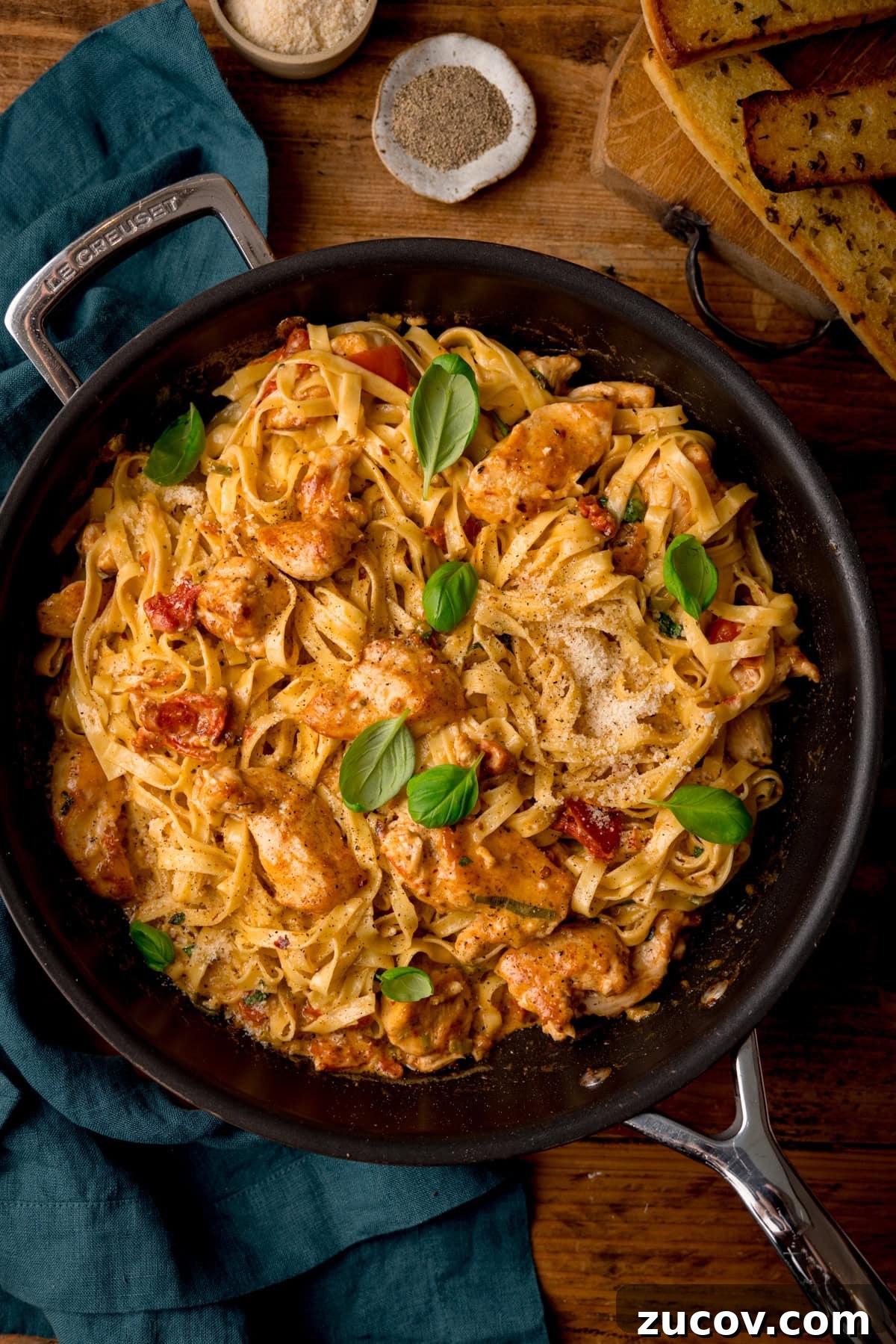 Creamy Marry Me Chicken Pasta Recipe for Weeknight Dinners 2 A tall, overhead shot of Marry Me Chicken Pasta in a black pan. To the left of the pan, there is a teal napkin tucked beside it. In the top right corner of the image, you can see a few slices of garlic bread in a stack, and to the left of this there are a few small dishes with seasoning, This is all set on a wooden surface.