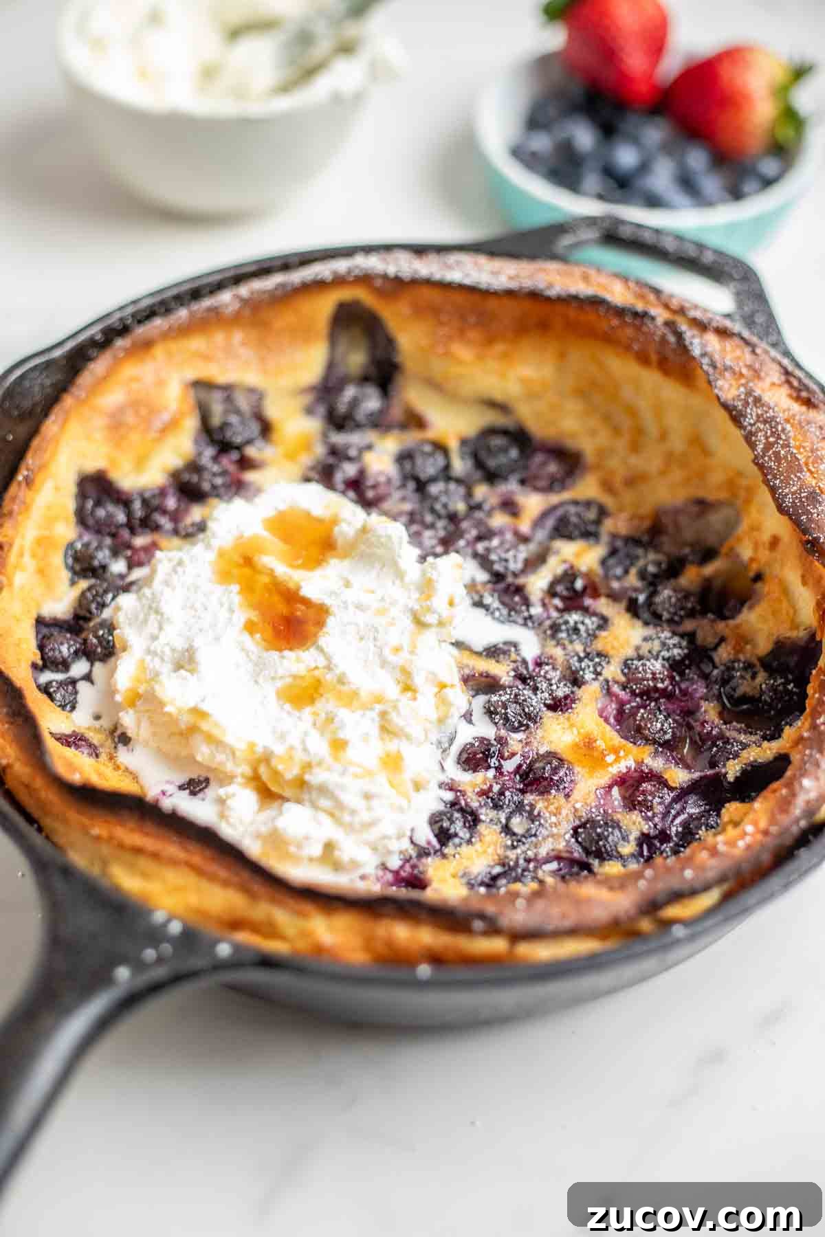 Blueberry Dutch Baby Pancake Recipe for Brunch and Dessert 2 Blueberry dutch baby topped with whipped cream and maple syrup in a cast iron skillet. A bowl of whipped cream and a small container of blueberries and raspberries are behind the skillet.