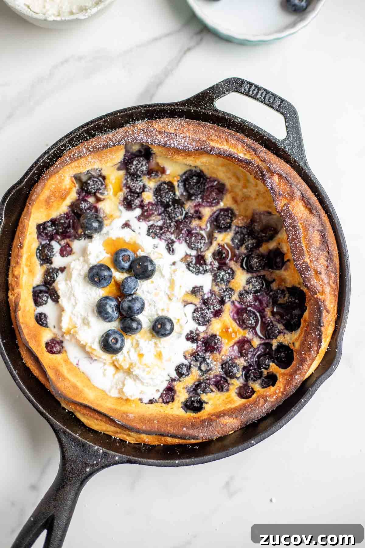 Blueberry Dutch Baby Pancake Recipe for Brunch and Dessert 9 Blueberry dutch baby pancake topped with whipped cream, blueberries, maple syrup and powdered sugar in a cast iron skillet.