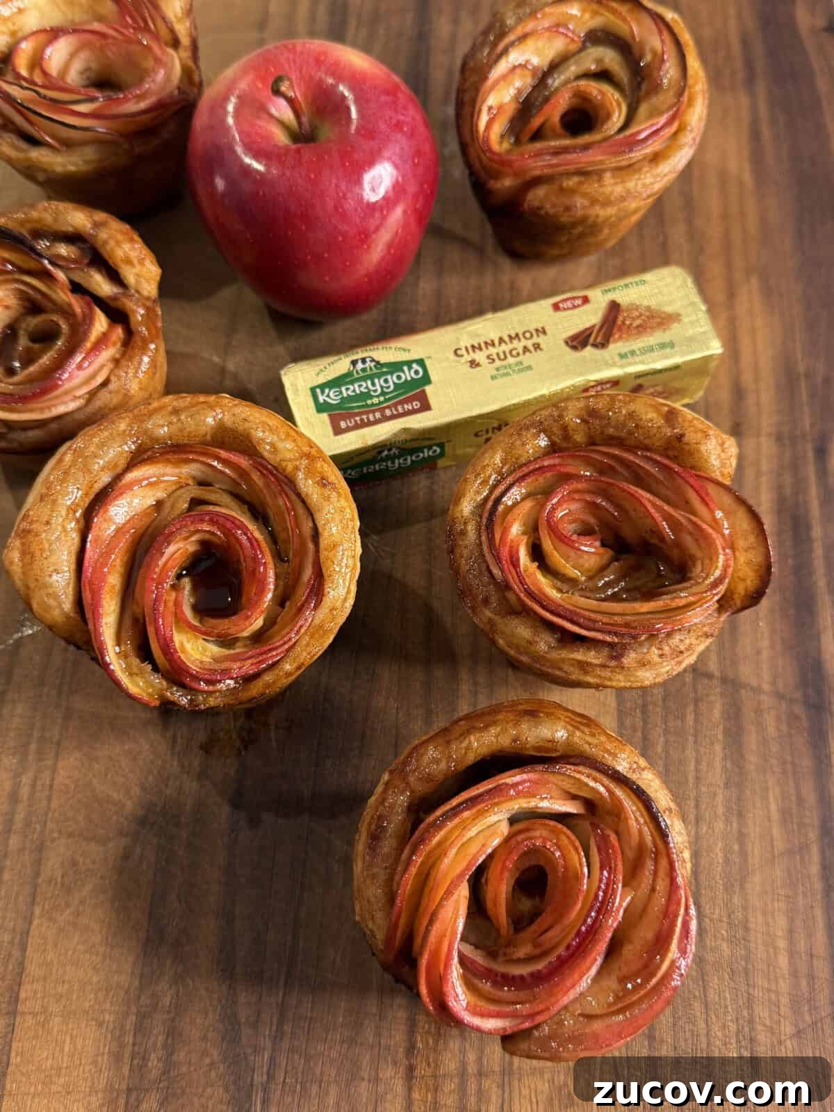 Apple Rose Pastry Recipe: Step-by-Step Homemade Apple Roses 2 Apple rose pastries are arranged on a wooden surface with a red apple and a pack of Kerrygold Cinnamon & Sugar Butter in the background. The pastries have thinly sliced apples forming a rose shape.